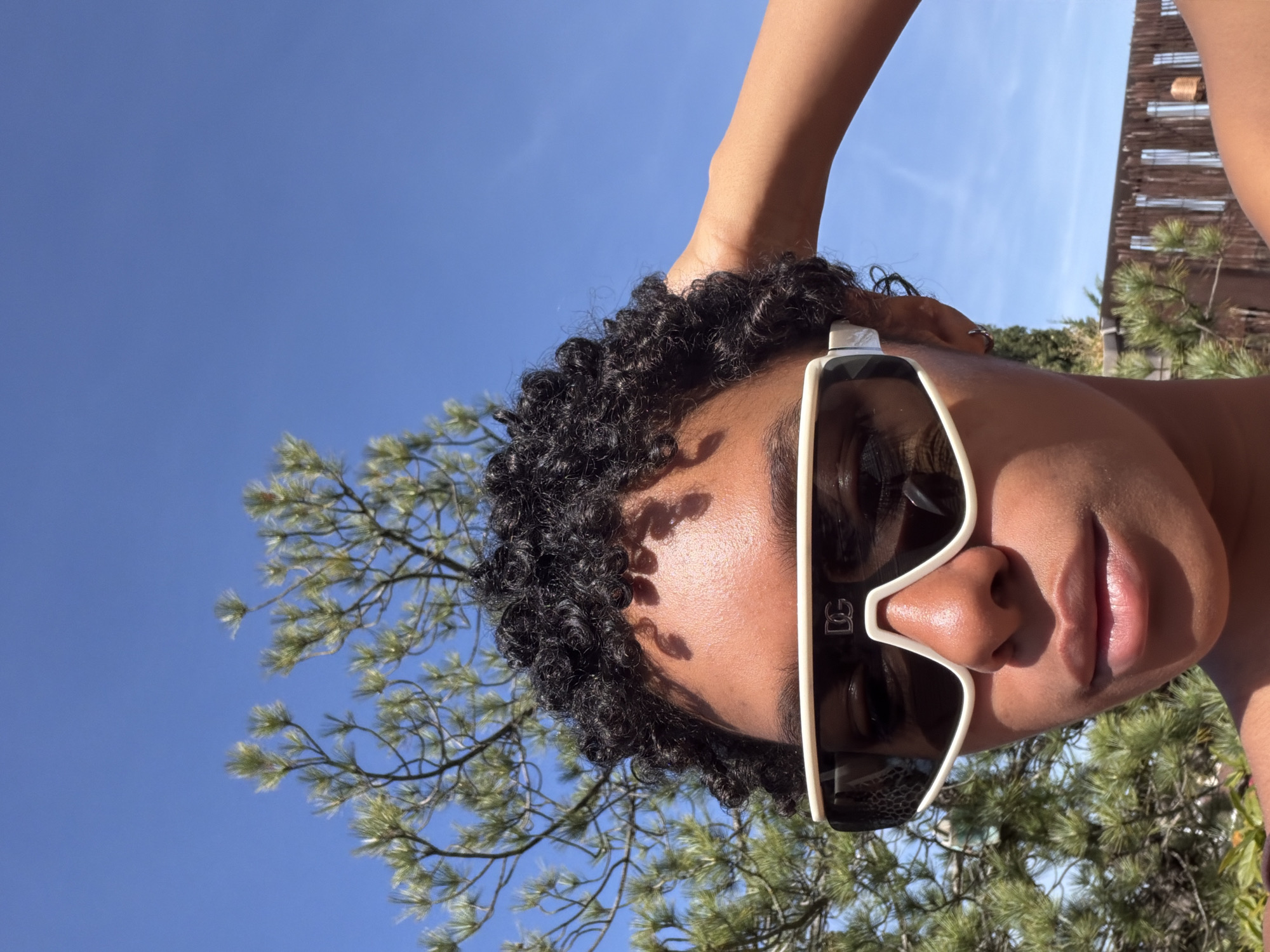 A low-angle, portrait-oriented photograph depicts a person with dark curly hair wearing sunglasses with white frames. The person's right hand is raised and appears to be touching their hair. The background consists of a clear blue sky partially obscured by the branches of a pine tree. A building with a wooden facade is also visible in the background. The photograph was not taken in Champs-sur-Marne, France.