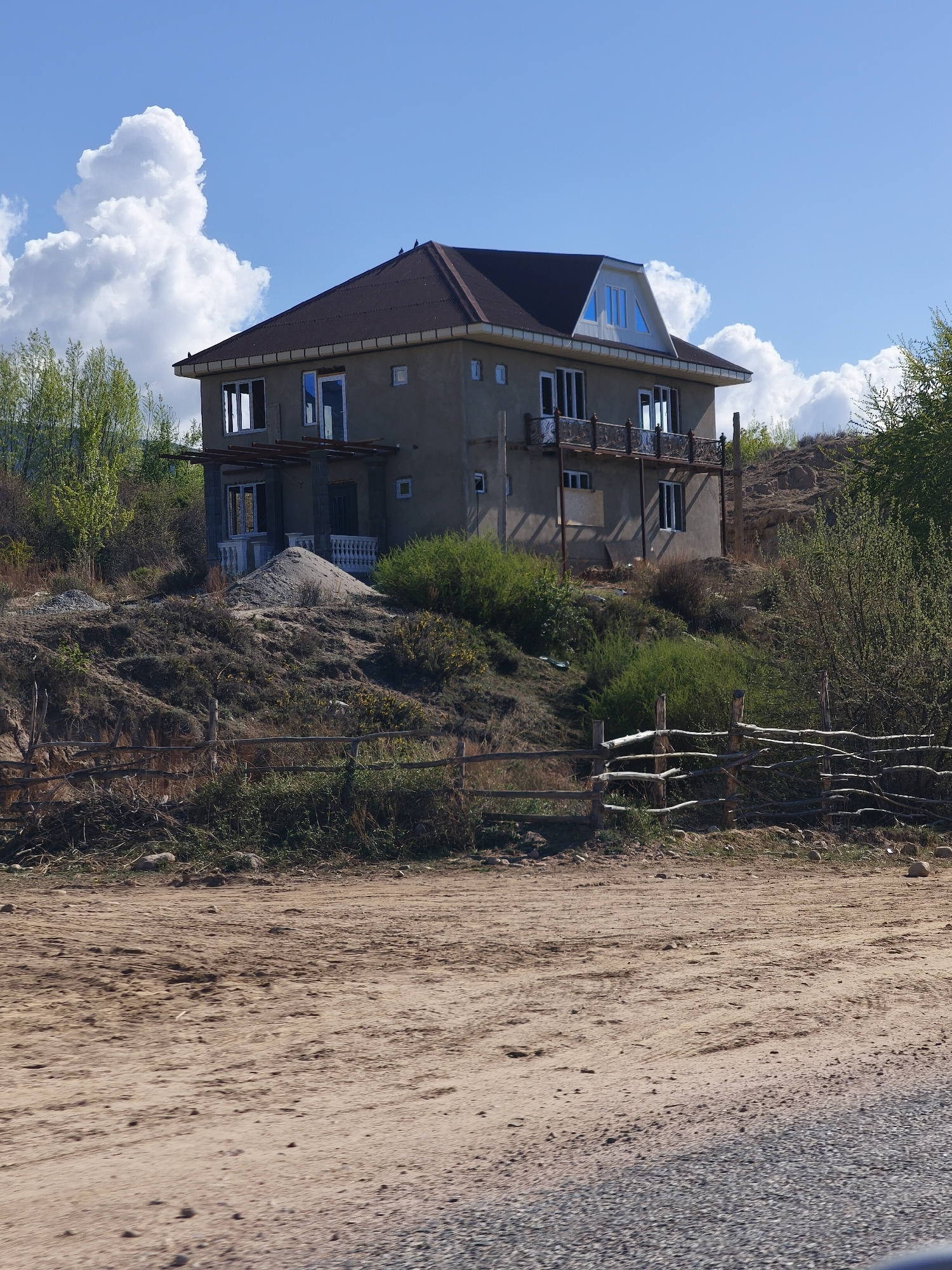 A wide-angle, daylight shot captures a two-story house situated on a hill under a bright blue sky dotted with large, fluffy white cumulus clouds.
In the foreground, a wide, sandy dirt road with visible tire tracks and scattered small rocks stretches across the bottom of the frame, transitioning into a gravel surface on the far right. A rustic wooden fence, constructed from irregularly shaped branches and logs, runs horizontally across the midground, delineating the road from a rising, uneven slope.
The house, the central subject, features a dark brown hip roof with light-colored eaves and neutral grey or beige stucco-like walls. It has multiple windows, some with white frames, and several balconies with decorative dark metal railings. A distinct dormer window with blue-tinted panes is visible on the upper roof section. On the lower level, a pile of light-colored sand sits next to what appears to be a covered porch area, suggesting ongoing construction or landscaping.
The terrain around the house is hilly and somewhat arid, covered with dry grass, sparse green bushes, and some taller, vibrant green trees, particularly on the left and right sides of the slope. The scene is brightly illuminated by the sun, indicating a clear day. No people or visible text are present in the image. The overall setting appears to be a rural or semi-rural landscape, consistent with the location context of Kiçi-Jargılçak, Kyrgyzstan.