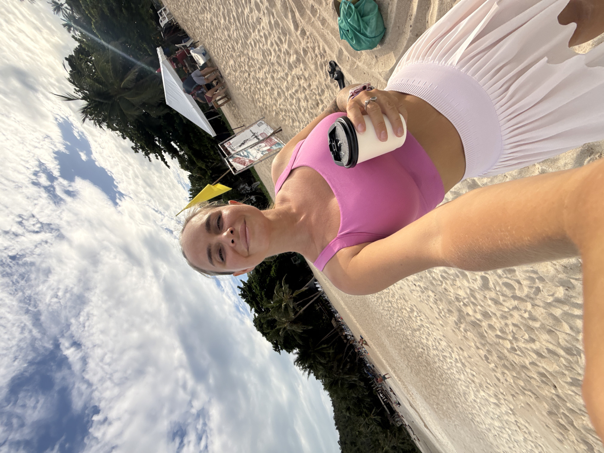 A woman stands on a beach in the Ko Samui District of Thailand, holding a coffee cup in her right hand. She is wearing a pink sports bra and a light pink skirt. The beach is sandy, and palm trees line the background. In the distance, a white tent is set up, with several people seated underneath it. The sky is partly cloudy.