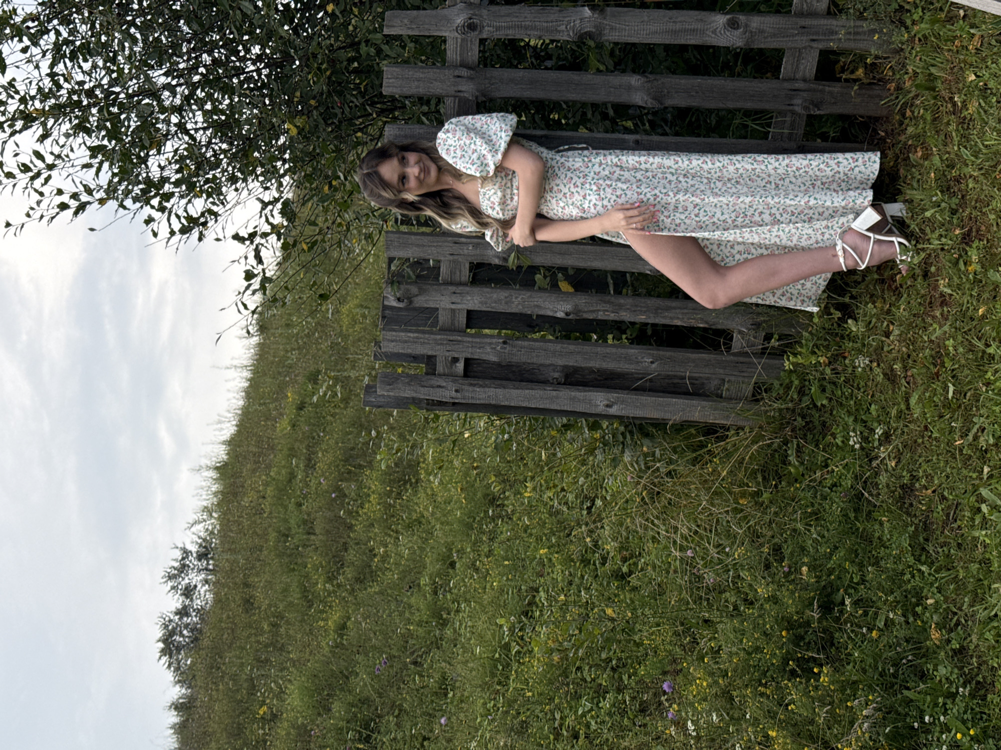 A young woman with long, blonde hair stands near a weathered wooden fence in a grassy, rural setting. She is wearing a white, floral-print maxi dress with puffed sleeves and a high slit on the side, revealing her legs and white, strappy heeled sandals. The woman is looking off to her left, with a slight smile. Behind her, a lush, green hillside is covered in a variety of wildflowers and grasses. A large tree with dark green foliage stands to her right, partially obscuring the sky, which is overcast and cloudy. The location is the village of Olkhovka, Russia.