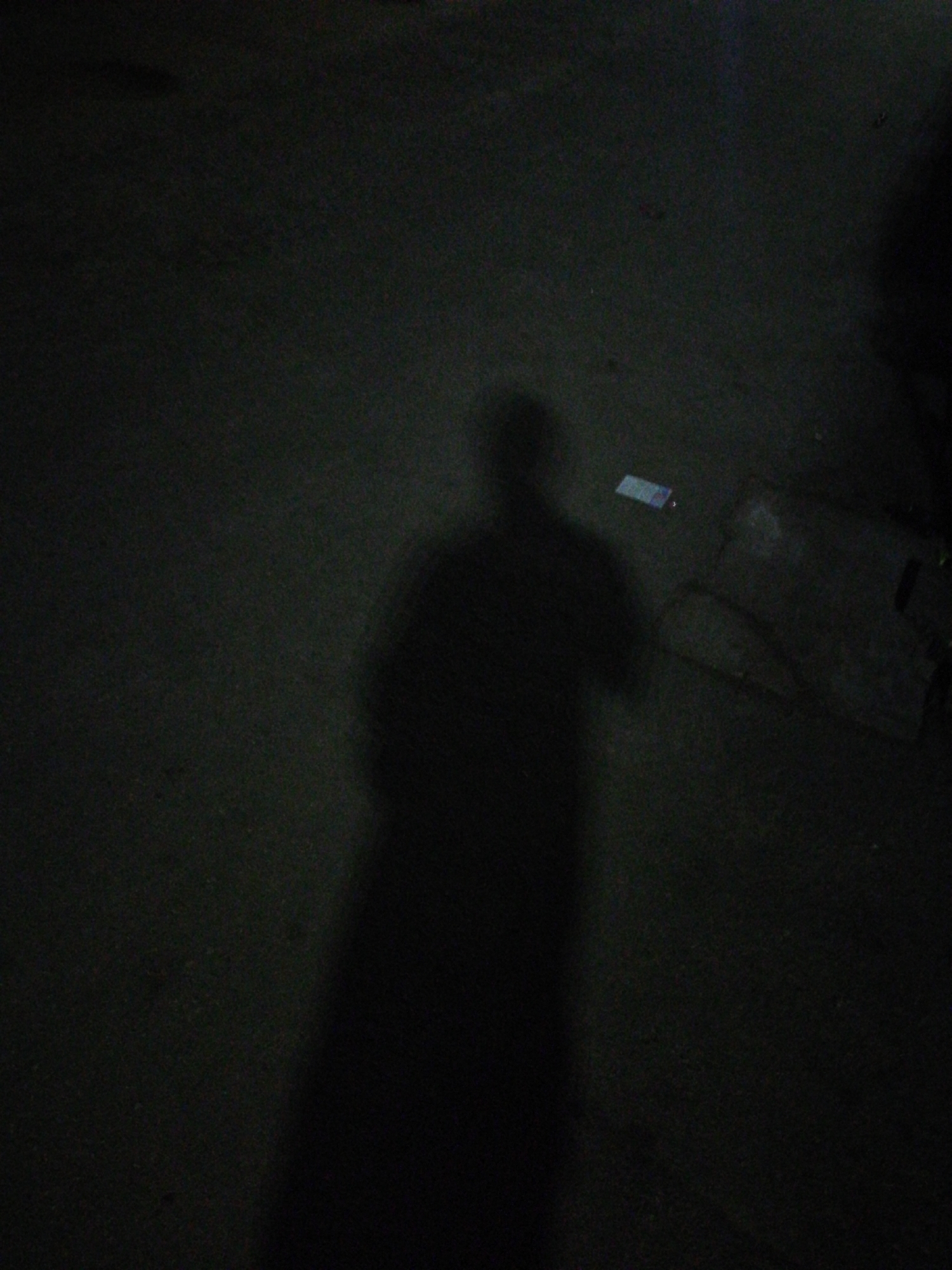The image is taken at night in Lagos, Nigeria. A large, indistinct shadow dominates the composition, suggesting a person illuminated by a light source behind them. A small, rectangular object, possibly a piece of paper or a similar item, lies on the ground nearby. The surrounding area appears to be paved, and a dark, possibly concrete structure is visible in the upper right quadrant of the image. The overall scene is dark with low visibility.