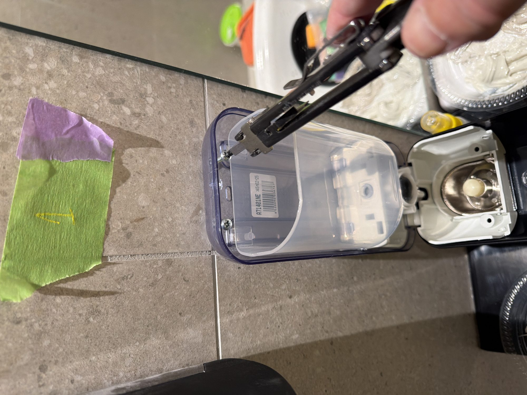 An outdoor scene shows a hand holding a pair of pliers, reaching towards a clear plastic dispenser mounted on a tiled wall. The dispenser has a label with the text "A71401NE X45H2125". Green and purple tape are affixed to the tiles above the dispenser, with a yellow mark resembling the number "1" on the green tape. The dispenser is open, revealing its internal components. In the background, a sink and other bathroom fixtures are visible. The overall setting appears to be a bathroom or a similar utilitarian space.