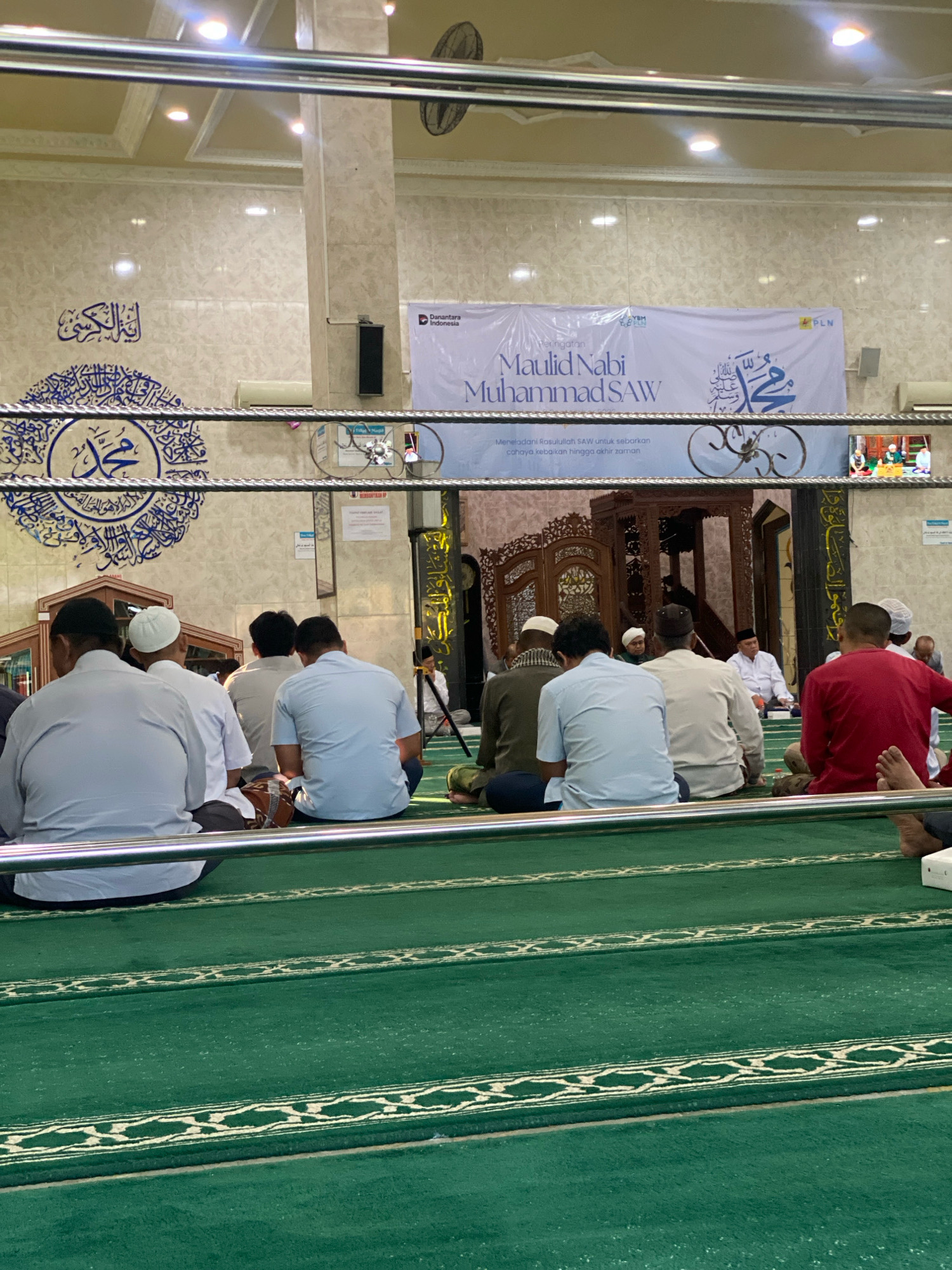The image depicts a gathering of men in a mosque in Banjarbaru, Indonesia, likely attending a religious event. The men are seated on a green carpeted floor, facing a decorated wooden structure, possibly a pulpit or mihrab. Some are wearing white religious caps, and others are in light-colored shirts. In the background, a banner with Arabic calligraphy and text that appears to be in Indonesian announces "Maulid Nabi Muhammad SAW," indicating a celebration of the Prophet Muhammad's birthday. The mosque interior features marble-patterned walls, intricate decorative elements, and lighting fixtures on the ceiling. The scene suggests a solemn and communal atmosphere.