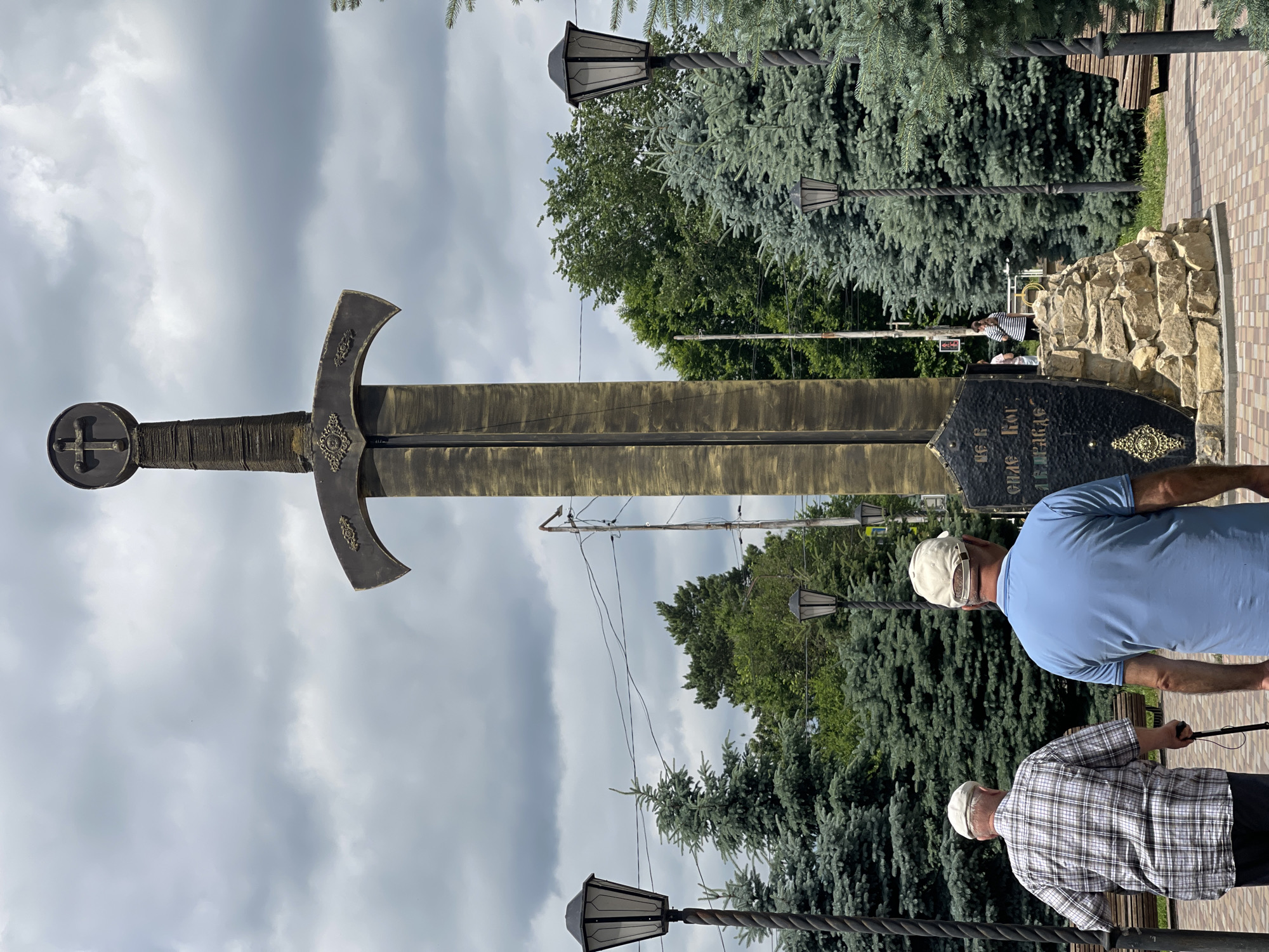 Two elderly men, one wearing a plaid shirt and the other a blue t-shirt, walk away from the camera towards a large sword monument in the city of Aleksandrovskoye, Russia. The monument, which appears to be made of metal, features a cross at its pommel and ornate detailing on the guard. It is mounted on a stone base. The scene is set outdoors with a cloudy sky above. Evergreen trees surround the monument, and lampposts are visible along the path. A small group of people is standing in the background near the monument. The ground is paved with interlocking bricks.