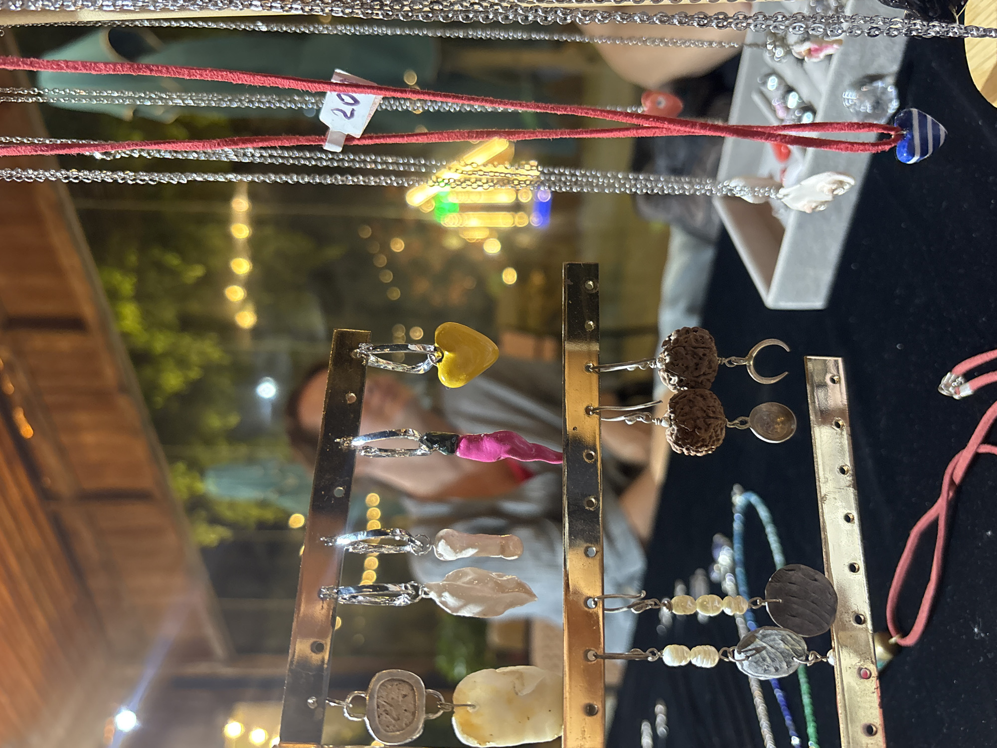 A close-up, eye-level shot captures an outdoor stall displaying various handcrafted jewelry items. In the foreground, a gold-colored rack holds an assortment of earrings. These include drop earrings with round, textured brown beads and crescent moon charms, pearl and disc drop earrings, and hoop earrings with various charms, such as a yellow heart, a pink and green object, and natural stone pieces. To the right, a collection of necklaces hangs. These are primarily silver chains, with one prominent red cord necklace adorned with a blue and white striped heart pendant. A small white tag with the number "20" is attached to one of the silver necklaces. In the background, a blurred human figure is visible, seated behind the stall. The environment appears to be a marketplace at dusk or night, suggested by the warm, out-of-focus bokeh lights in the background, indicative of ambient lighting. The stall itself is under a wooden overhang, with greenery visible beyond.
