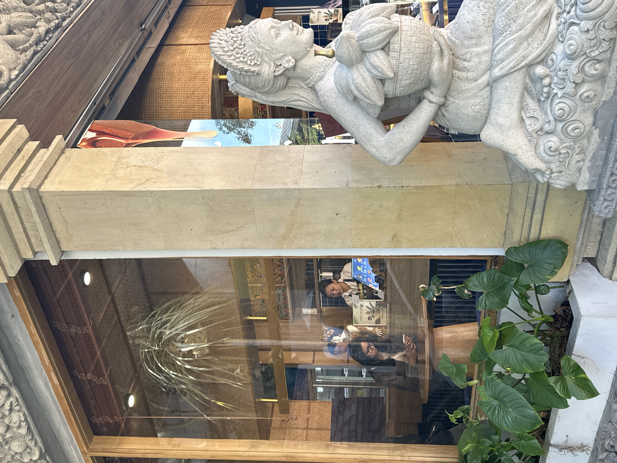 In front of a cafe in Ubud, Indonesia, a stone statue of a woman with an elaborate headdress and a basket of fruits stands to the right of the frame. Through the cafe's large window, two women are visible. One woman sits at a circular wooden table, looking at her phone, while another woman stands behind a counter, appearing to be an employee. The cafe's interior features wooden accents, woven wall coverings, and a hanging plant. Lush green plants in a planter box are in the foreground to the left of the statue.