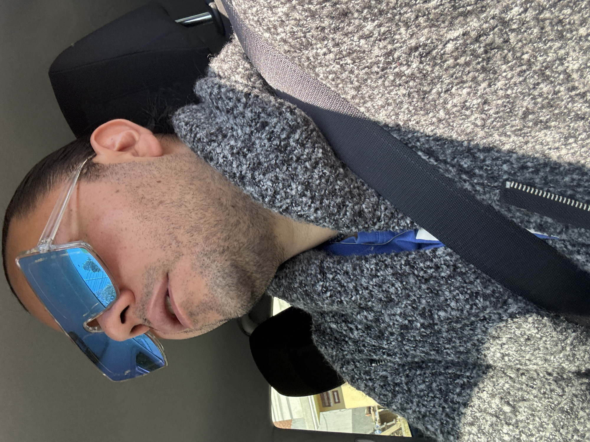 A male individual is observed seated in the front passenger compartment of a vehicle in San Juan Cuautlancingo, Mexico. He wears large, clear-framed sunglasses with reflective blue lenses, which show reflections of power lines and tree foliage. The subject has short facial hair, and his dark hair appears pulled back. He is dressed in a grey, textured, collared sweater with a visible zipper closure, layered over a blue garment. A black seatbelt is fastened across his chest and left shoulder. The vehicle's interior features a black headrest. Outside the vehicle, visible through a side window, is a light-colored building with a distinct yellow section.