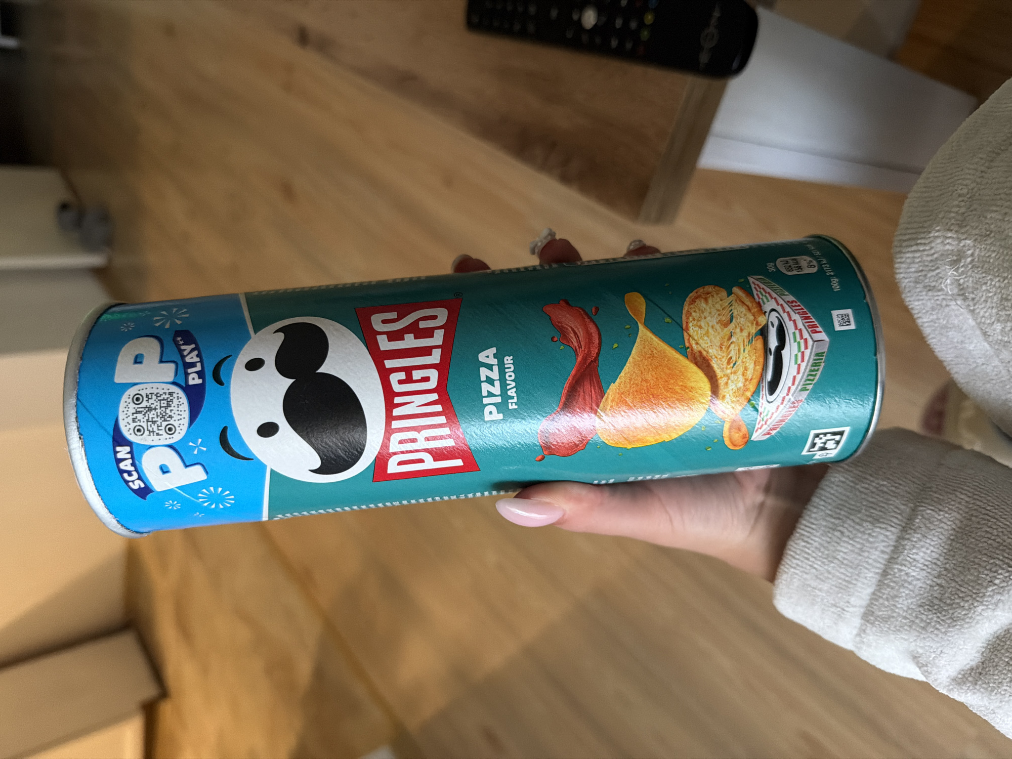 The image depicts a Pringles can held by a human hand, with the scene reportedly located in Plovdiv, Bulgaria. The Pringles can, oriented vertically and slightly angled towards the left, is "Pizza Flavour" and features the brand's iconic Mr. Pringle character. The top section of the can is blue with "SCAN POP PLAY" text and a visible QR code. The main body is teal, displaying the red "PRINGLES" logo, "PIZZA FLAVOUR" text, and an illustration of potato chips, melted cheese, and red sauce. A graphic resembling a pizza box, labeled "PRINGLES PIZZERIA," is present near the bottom. Nutritional information, including "100g: 2173 kJ / 521 kcal," is partially visible on the lower right. The can's foil seal is evident at its rim.
A hand with light skin tone and manicured, light pink/nude oval-shaped nails firmly grips the can. The individual's wrist and lower forearm are covered by a light-colored, possibly off-white, terry cloth material, suggesting comfortable loungewear.
The background shows an indoor environment with light-colored wooden floorboards, likely laminate or parquet, extending into the distance. To the right, a dark, rectangular remote control, consistent with a television remote, rests on a light-colored wooden surface, possibly a table or shelf. In the far background, light-toned walls are visible, along with indistinct furniture or architectural features, suggesting a domestic setting such as a living room. The background is moderately out of focus, emphasizing the can and hand.