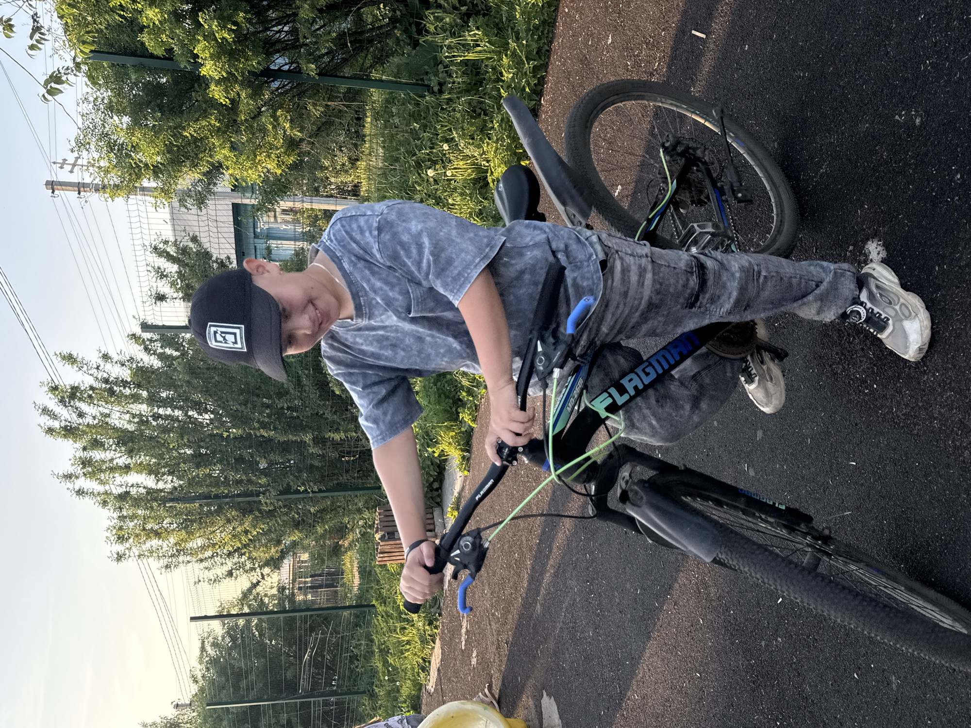 A young person stands on a bicycle on an asphalt surface. The person wears a dark baseball cap, a patterned gray t-shirt, and gray pants. The bicycle has black and blue accents, with the word "FLAGMAN" visible on the frame. The setting appears to be in an urban area in the city of Фоки, Russia, as indicated by the buildings, fence, and vegetation in the background. The weather is bright and sunny.