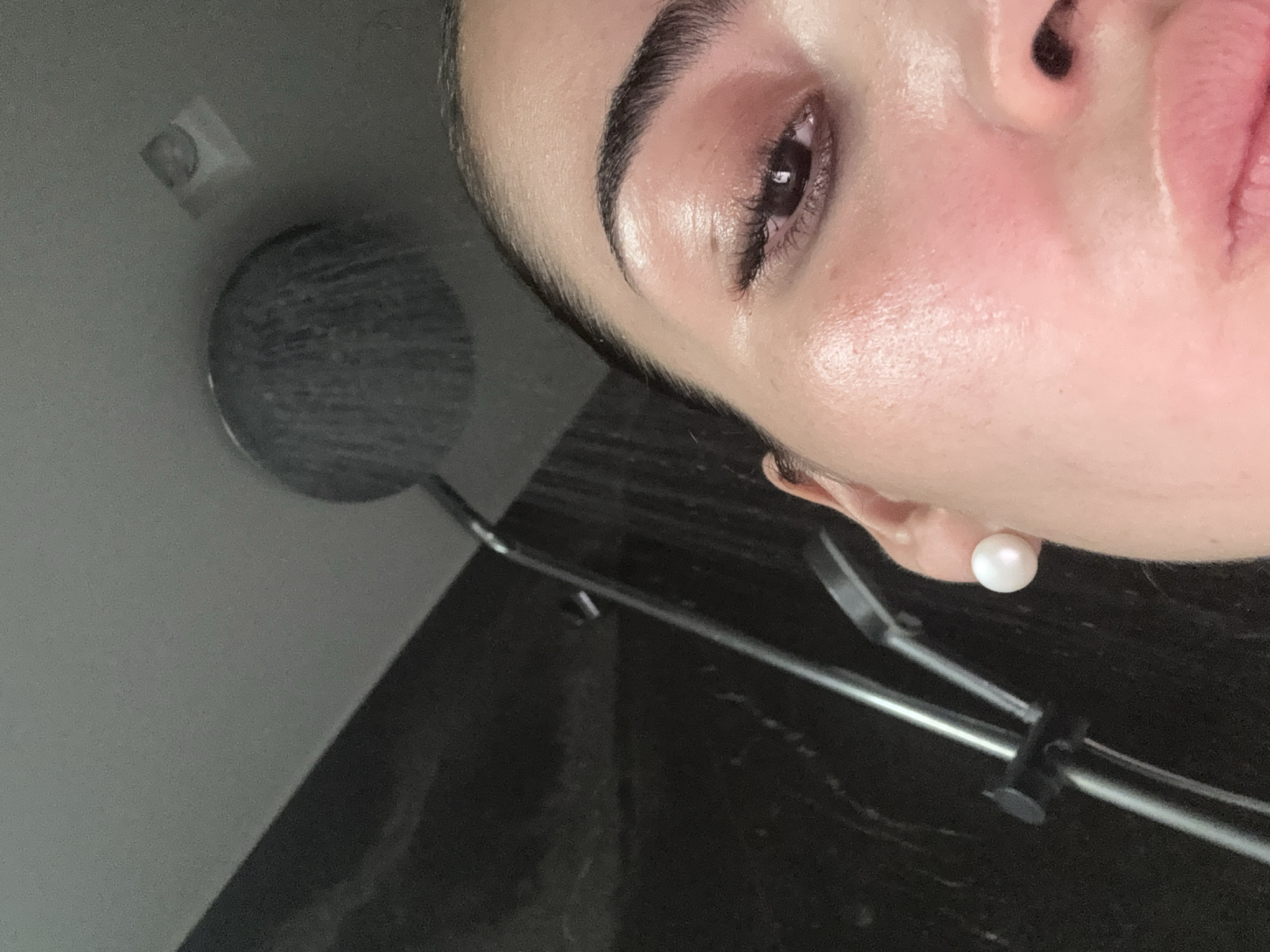 A close-up photograph shows a woman's face, turned to the right, with a showerhead visible on the left. She has dark eyebrows, eyelashes, and a pearl earring. Her skin is fair. The showerhead is a circular, modern design. The background is composed of gray and dark marble-like surfaces. The photograph provides no information linking it to San Bernardino Tlaxcalancingo, Mexico.