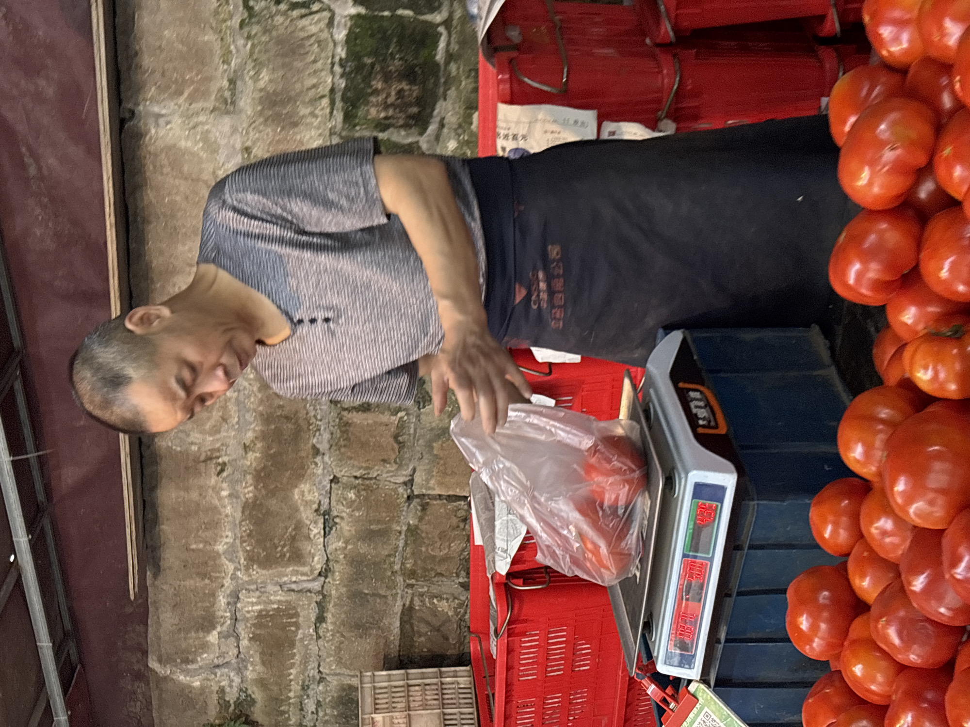 An Asian man is weighing tomatoes on a digital scale at an outdoor market. He wears a short-sleeved, striped shirt and a dark apron. The scale displays the weight of the tomatoes in a plastic bag. A large pile of red tomatoes is next to the scale. The market appears to be in the city of Jiangjin, China, based on labels in the image. The backdrop features a brick wall.