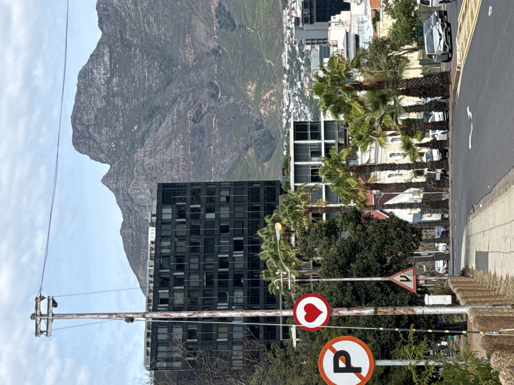 The scene is in Cape Town, South Africa, looking towards Table Mountain. The view includes a tall, dark building with many windows, palm trees, and a road with cars and other buildings. Street signs are visible in the foreground, including a heart symbol and a no-parking sign. An electricity pole with wires is on the left side of the image.