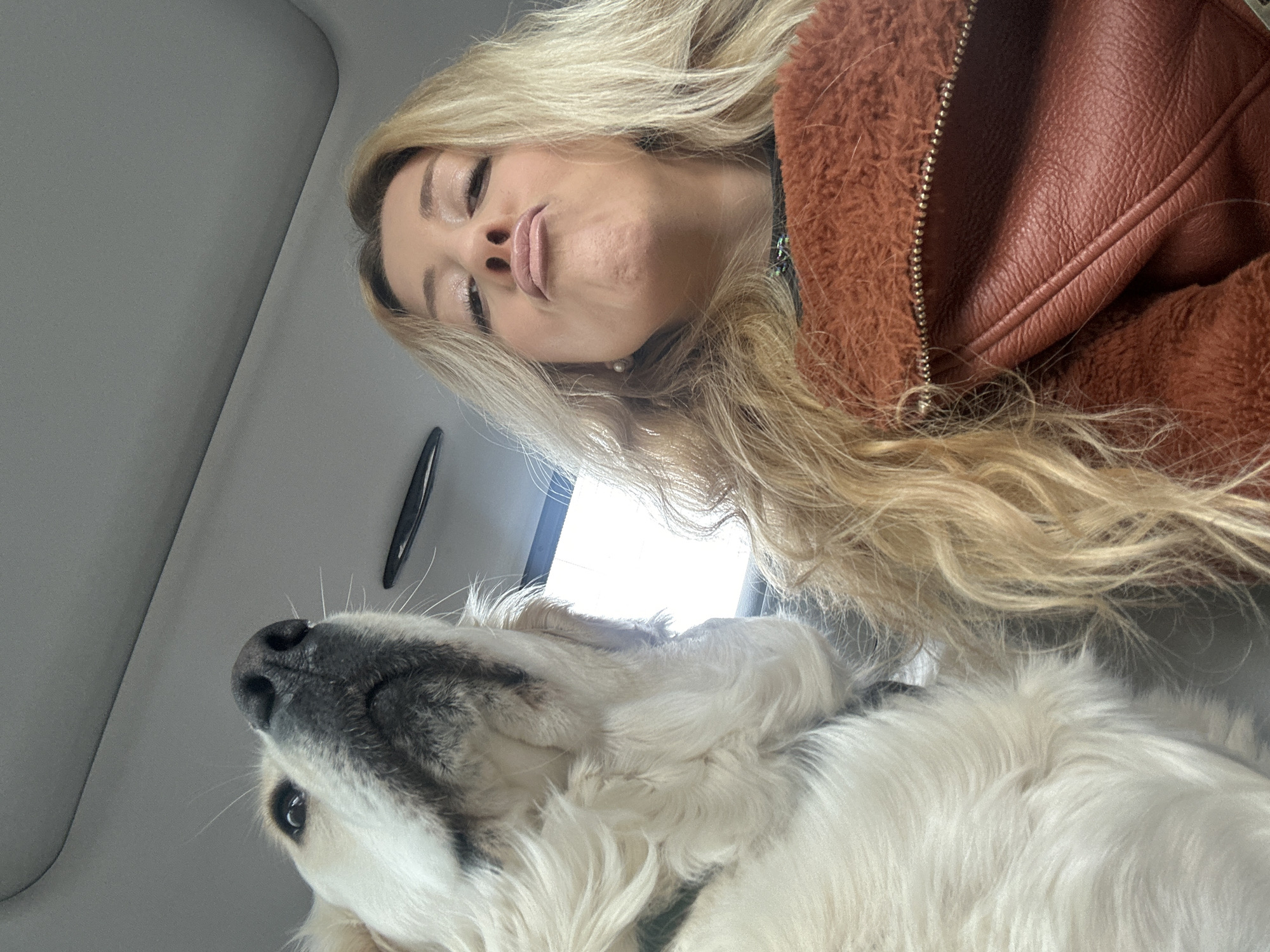 A selfie taken from inside a car shows a blonde woman with puckered lips looking down at a white dog with a black nose. The car's interior is visible, with a light gray ceiling and a dark handle. The woman is wearing a brown jacket with a furry collar. The dog is looking up at the woman. The setting is implied to be in a car, but there are no specifics of the city of London.
