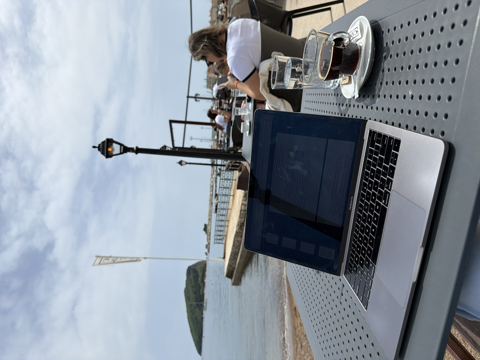 The photograph captures a scene outdoors at a cafe or restaurant, likely located in or near Budva, Montenegro, given the presence of an island in the background and the setting that appears to be along the coast. The focus is on a metal table featuring a laptop computer, a cup of coffee, and a glass of water. The laptop is open, suggesting someone may be working or browsing. A person is visible in the background, seated at a table. Other patrons may be at tables in the far background. The environment includes a waterfront area with a pedestrian walkway, lampposts, and a glimpse of the sea with a small island in the distance. The sky is visible above, partially obscured by the cafe structure and foliage.