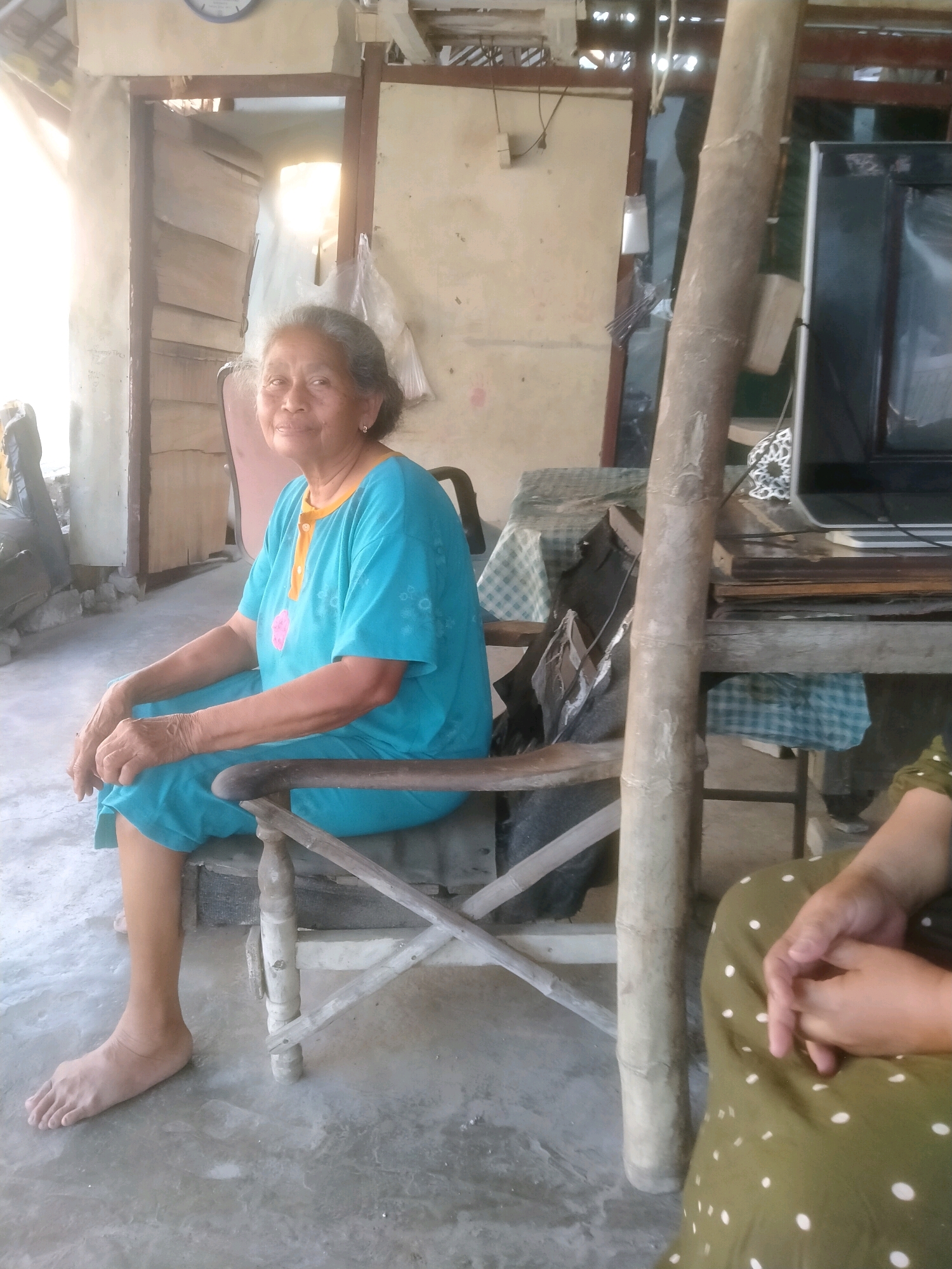 An elderly Indonesian woman with gray hair, wearing a turquoise t-shirt and pants, sits on a wooden chair outdoors in what appears to be a rustic setting. Sunlight illuminates the scene from the left. Behind her, a doorway leads to a dimly lit interior with wooden walls. A large, rough tree trunk stands prominently in the foreground, leaning towards the right. To the right of the tree trunk, another person's hands are visible, clasped, resting on a green, polka-dotted fabric. A table with a checkered cloth is present, and part of a television set is visible on the right side of the frame. The ground is concrete. The image suggests a casual, everyday moment in a rural or semi-rural Indonesian environment, possibly in Sumengko, Indonesia, given the context.
