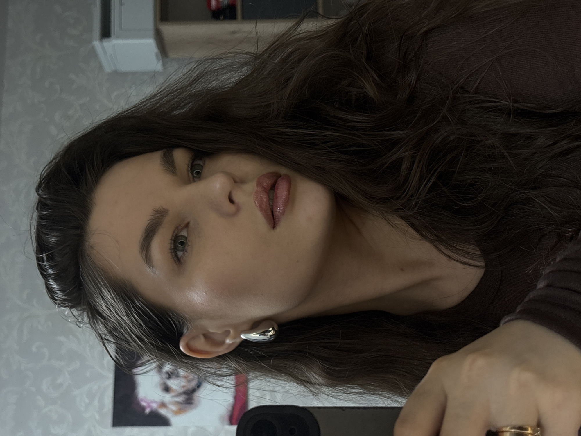 The image is a close-up, selfie-style portrait of a young woman with long, dark, wavy hair. She is wearing large, silver, teardrop-shaped earrings and a brown top. Her makeup includes noticeable lip gloss. The background appears to be an indoor setting with a patterned wallpaper and a framed picture of a small white dog visible on the left. On the right, a shelf unit with some items is partially visible. The lighting suggests an indoor environment, possibly a home. There is no indication of the city of Chaikovsky, Russia, in the image.