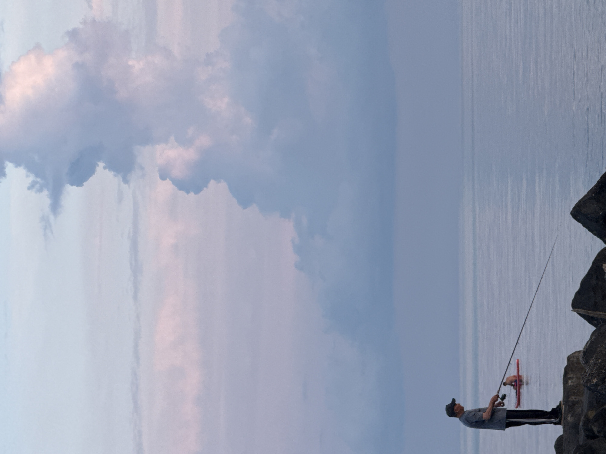 In Nebug, Russia, on a tranquil body of water, a lone fisherman stands on a rocky shore, casting a fishing line into the calm sea under a pastel-colored sky. The sky is a blend of soft blues, lavenders, and pinks, with large, ethereal clouds dominating the upper portion of the frame. In the distance, a smaller red float is visible, likely part of another fishing setup, with a person on it. The foreground features rugged, dark rocks, providing a stable platform for the fisherman. The overall atmosphere is peaceful and serene, with the soft hues of the sky reflecting on the still water.