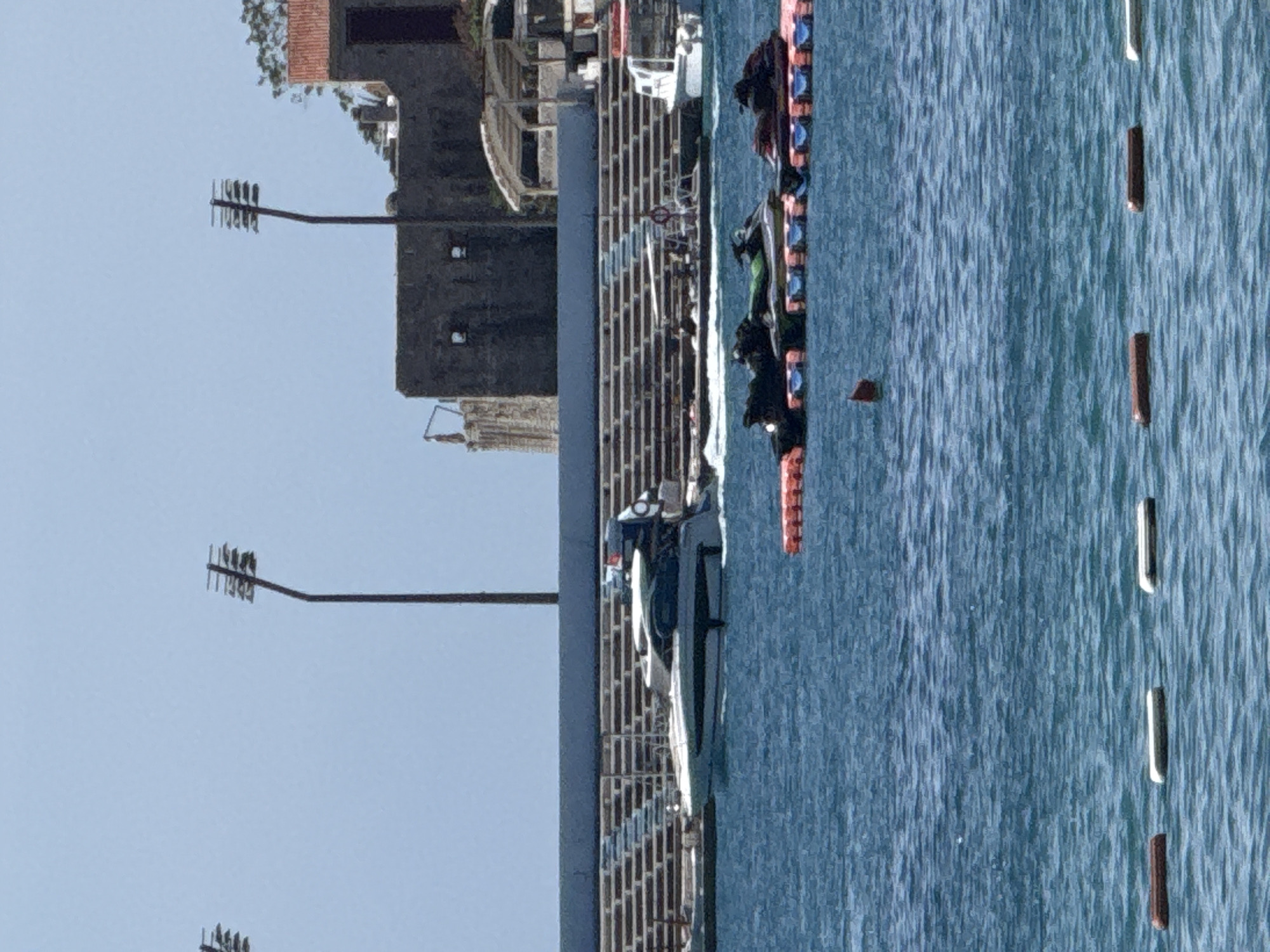 The image depicts a scene at the Budva waterfront, Montenegro. In the foreground, several dark-colored jet skis are docked on a floating platform in the blue water. Further out in the water, a white motor yacht is cruising, leaving a wake. Behind the yacht, a long, tiered seating structure, likely a stadium or amphitheater, is visible, bordered by a blue barrier. In the background, an old stone building complex with red-tiled roofs sits atop a hill. Two tall lamp posts, characteristic of stadium lighting, stand against a clear blue sky. The overall atmosphere suggests a sunny day at a recreational coastal area.