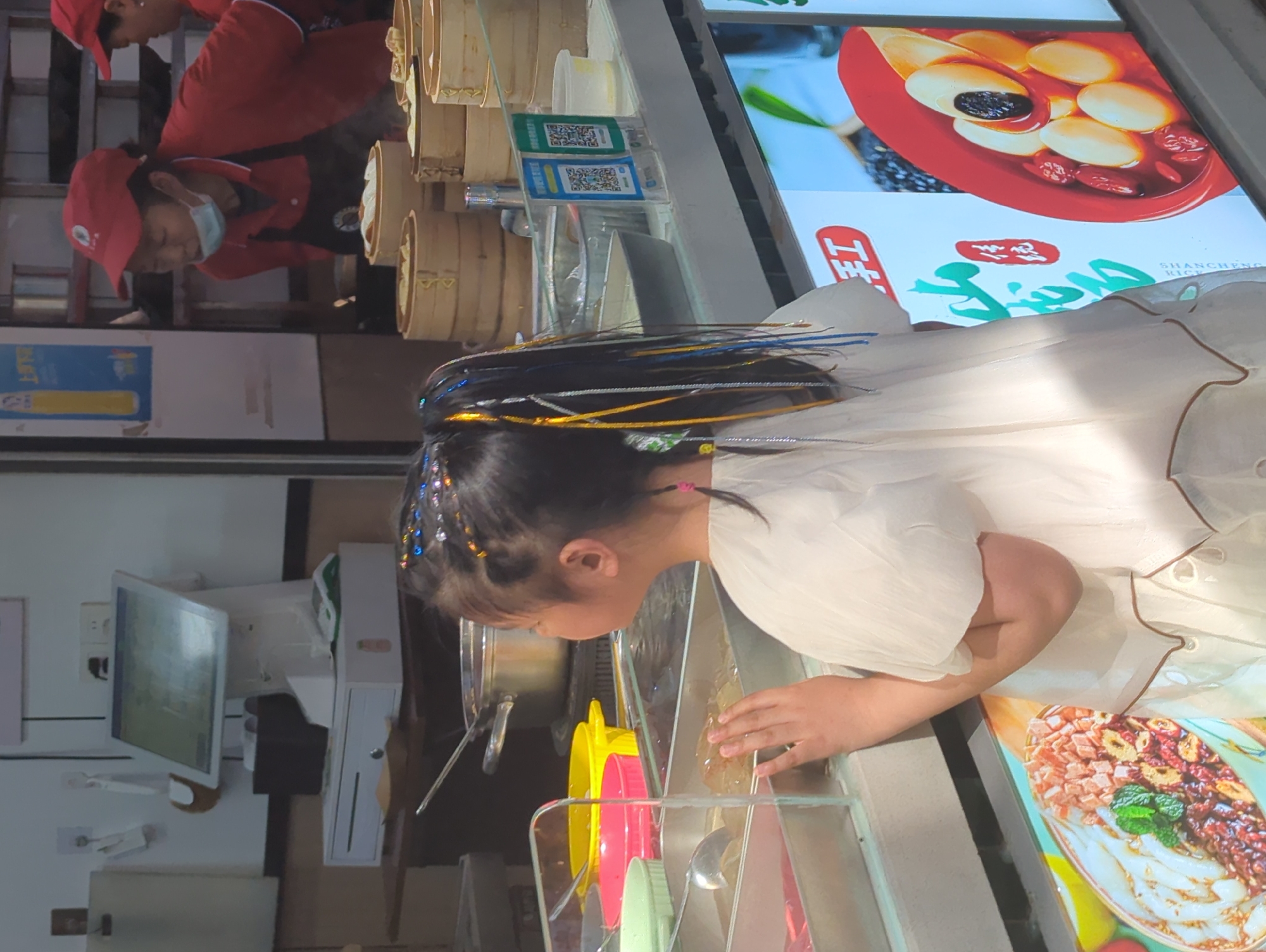 The image depicts an indoor scene within a food establishment, likely a dessert shop or food stall, located in Yuzhong District, China. In the foreground, a young girl, dressed in a white outfit, leans forward to peer into a glass display counter. Her dark hair is styled with numerous colorful beads and extensions in shades of blue, yellow, and silver. She appears focused and curious, with her hands resting near the glass, examining the contents. Inside the counter, various food items or toppings are visible in colorful bowls, including yellow, pink, and green.
Behind the counter, two staff members wearing red caps and red uniforms are present; one staff member also wears a light blue face mask. Above them, stacked bamboo food steamers suggest dishes are being prepared or kept warm. Payment options are displayed prominently on the counter with two QR codes, one green and one blue, indicating mobile payment methods. A large digital display screen showcases appetizing food imagery, including a red bowl containing a dessert with white and red ingredients, and another dish adorned with various toppings. Text visible on the screen includes "上品" (Shàngpǐn), "米" (Mǐ), and "SHANCHENG RICE," hinting at the type of cuisine served, possibly rice-based desserts or dishes. To the left, a computer monitor and a white cash register unit form part of the point-of-sale system. The scene is brightly lit, suggesting it is daytime or well-lit artificially, creating a lively atmosphere within the food venue.