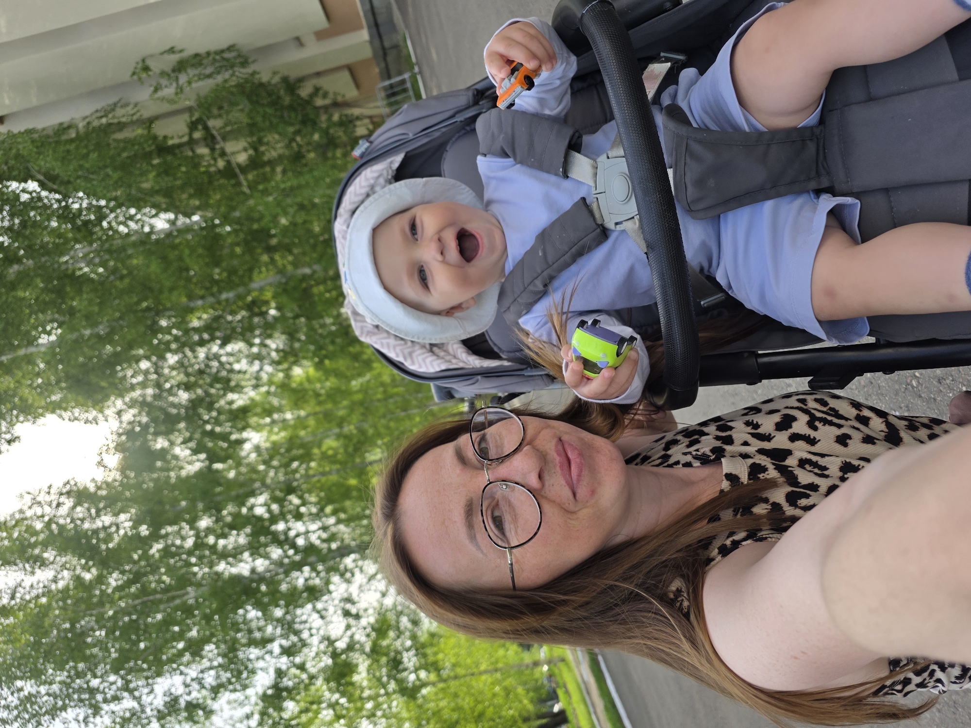 The image shows a selfie taken by a woman wearing glasses and a leopard-print top. In the background, a baby wearing a bonnet and a blue outfit sits in a gray stroller, holding small toy cars. The location appears to be an outdoor area in the city of Chaikovsky, Russia, based on contextual clues. The scene features foliage and urban structures in the background. The woman is holding the camera, capturing herself and the baby in the stroller.