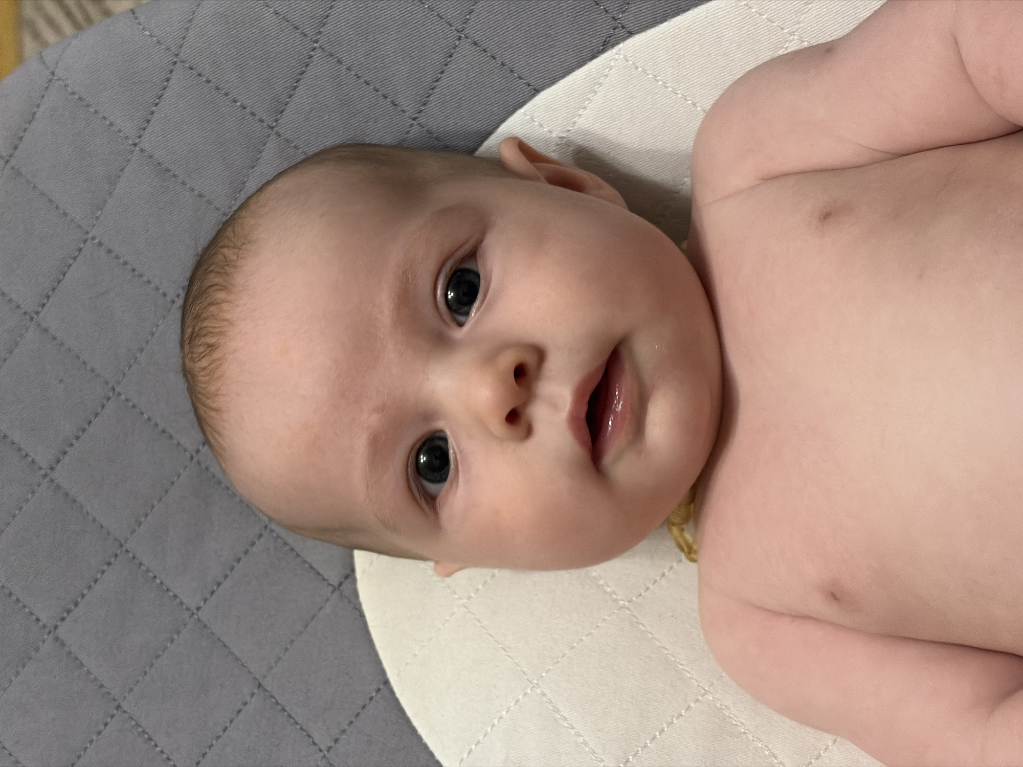 A baby with fair skin and dark eyes lies on a quilted surface. The surface is comprised of two sections, gray with a diamond pattern on the left and white with a similar diamond pattern on the right. The baby is positioned with its head to the left and its body extending to the right. The baby is naked. The location is not discernible from the image.