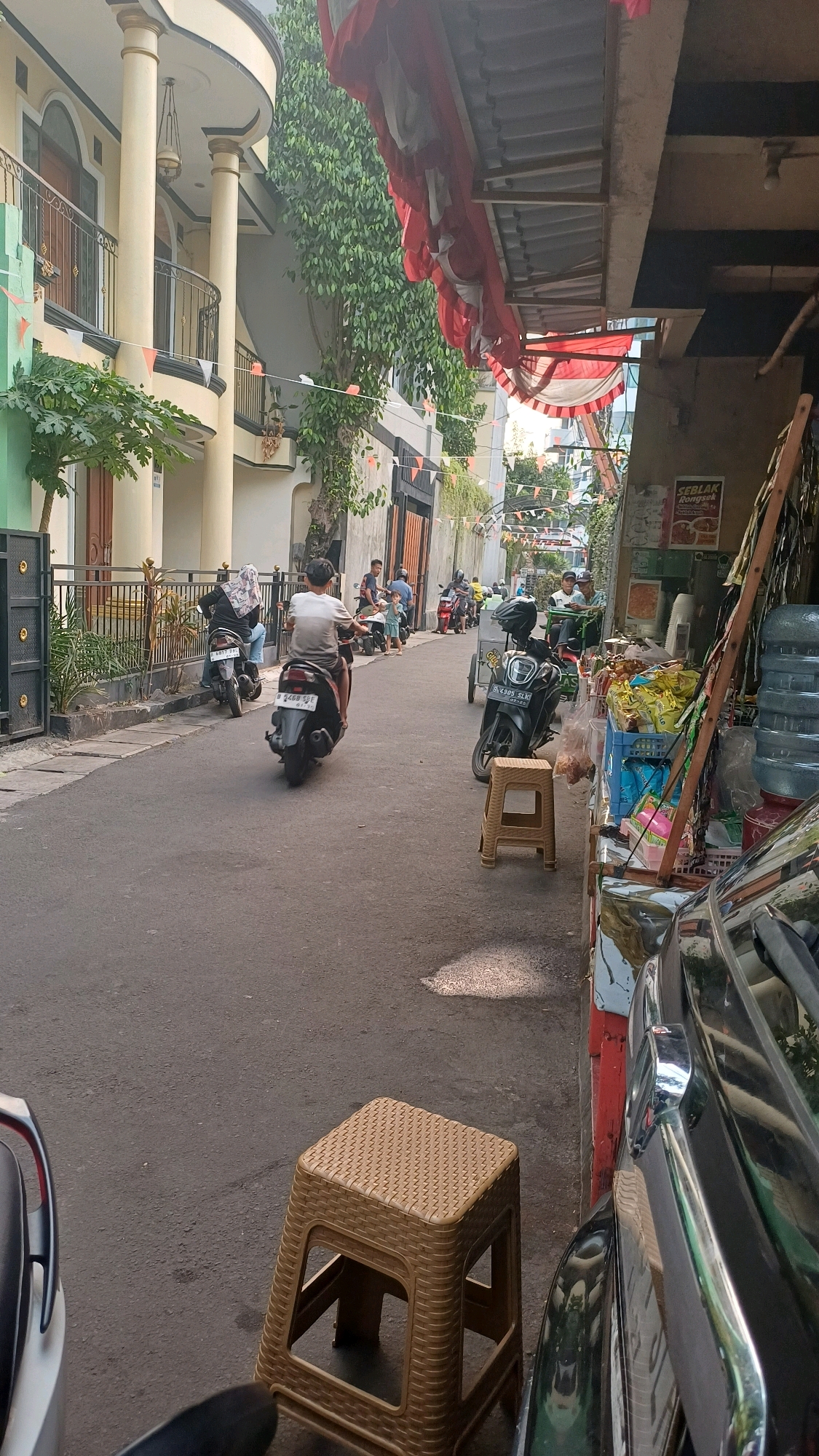 The image captures a narrow street in a residential area of South Jakarta, Indonesia, likely on a sunny day. The street is paved with asphalt and bordered by a sidewalk with stone paving. On the left, a light yellow multi-story building with white columns and balconies is visible, adorned with green plants. On the right, a street-side stall or shop is set up under a corrugated metal roof covered with red and white fabric decorations, reminiscent of Indonesian Independence Day.
Several motorcycles are present, with some being ridden. In the foreground, a person on a dark-colored scooter with a license plate beginning "B 2468 SJE" is moving away from the camera. Another scooter is parked on the left side of the street, with a person wearing a headscarf seated on it. Further down the street, more motorcycles and people are visible, including a person in a white shirt and dark shorts riding a scooter. The stall on the right displays various items, including bottled water, packaged snacks, and other goods. A light brown plastic stool is placed in front of the stall on the asphalt. A dark-colored car is partially visible on the far right, in front of the stall. The overall impression is a busy, everyday scene in an urban Indonesian neighborhood.