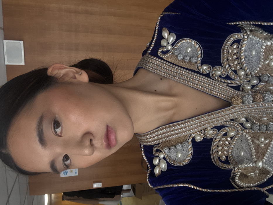 A close-up portrait depicts an individual with East Asian features wearing an ornate, deep blue velvet garment, heavily embroidered with gold, silver, and various pearl-like beads along the V-neckline and shoulders. The person has light makeup, dark hair pulled back into a bun, and a small stud earring in the right ear. Two distinct moles are present on the neck. The background consists of vertical wooden paneling, with a white rectangular fixture and a partially visible Wi-Fi symbol sticker. The scene is located indoors in Paris, France. The individual is looking directly at the camera.