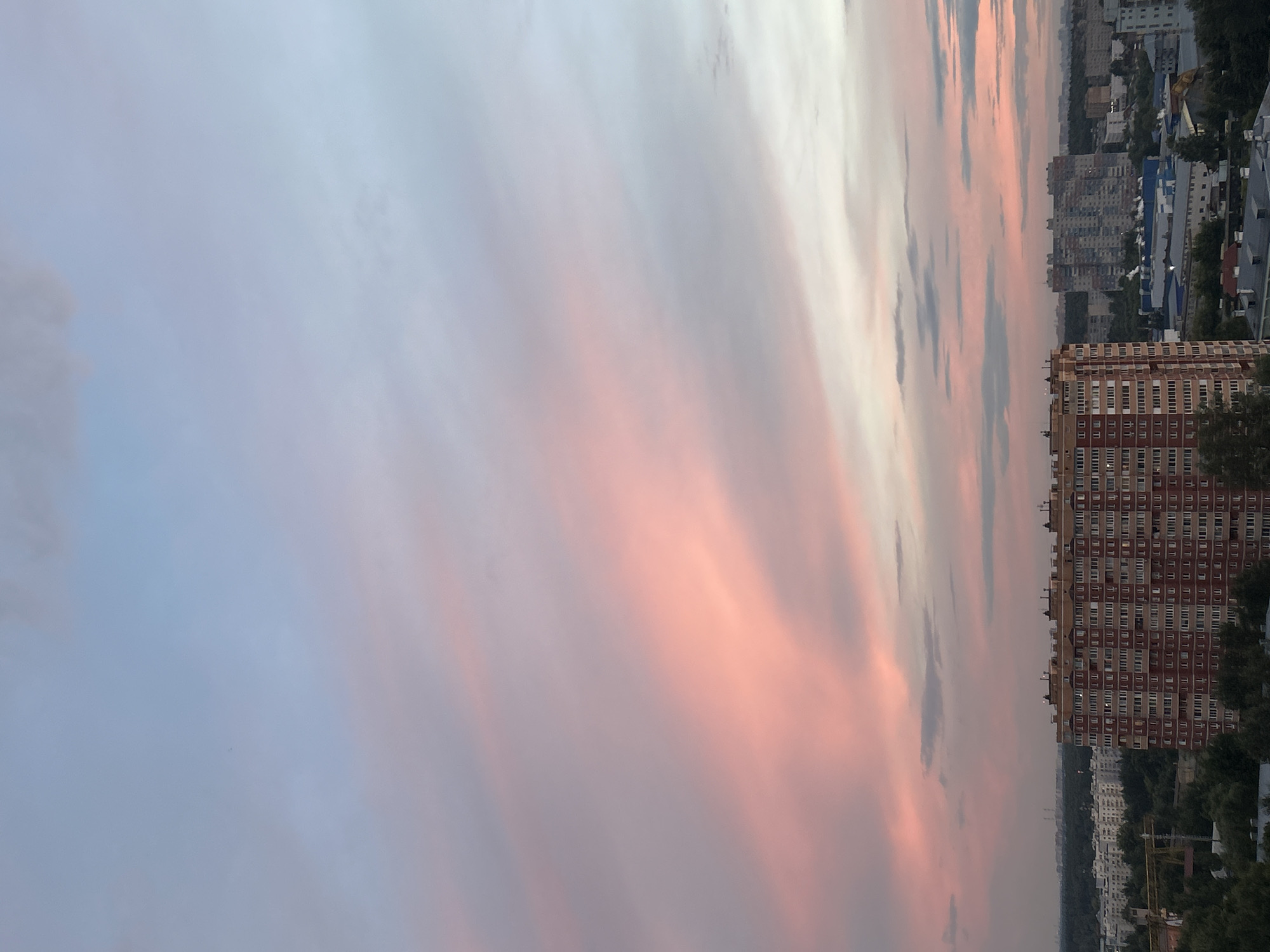 The image shows a cityscape at dusk, with a prominent red-brick apartment building in the foreground. The sky is filled with soft, pink and grey clouds, characteristic of a sunset or sunrise. In the background, other buildings and trees are visible, stretching towards the horizon. The overall scene suggests an urban environment in Ivanteyevka, Russia, during the twilight hours.