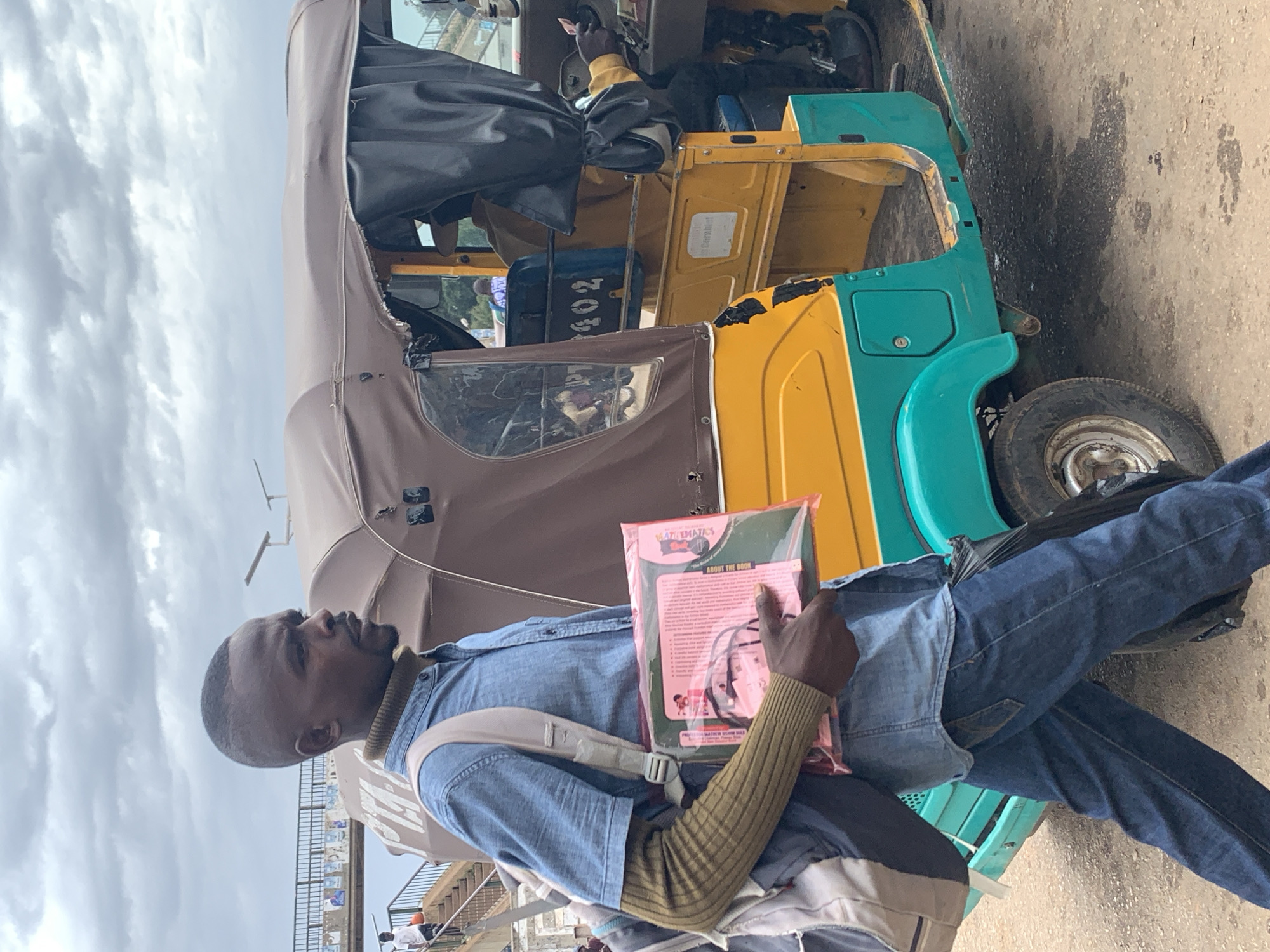 A man walks past a yellow and teal tricycle with a brown canopy in Gura Top, Nigeria. The man, with dark skin and a close-cropped haircut, wears a blue denim shirt over a green ribbed sweater, dark blue jeans, and a grey backpack. He carries a clear plastic-wrapped bundle of pink booklets in his right hand. The tricycle has a dark curtain on the right side and a number plate with "402" visible. The ground is a dirt and gravel surface, and the sky is overcast with grey clouds. In the background, a pedestrian bridge with railings and a few people can be seen, along with a lamppost.