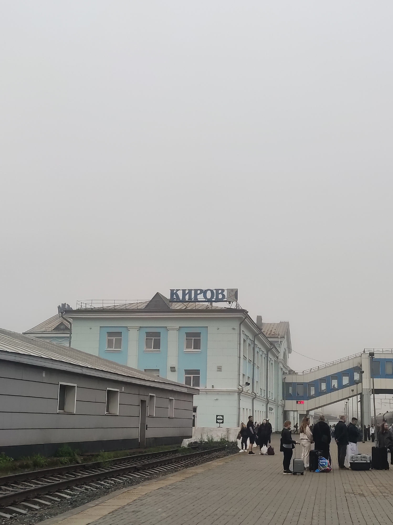 The image depicts a railway station in Kirov, Russia, on an overcast day. A two-story building with light blue and white accents, featuring multiple windows, is visible. The word "КИРОВ" (Kirov) is displayed in large blue letters on the roof of the building. To the left of the main building is a long, single-story grey structure with windows. A railway track runs in front of these structures.
On the platform in front of the main building, a group of people are standing, some with luggage, appearing to be waiting for a train. There is an elevated walkway connecting to the building, with windows along its length. A digital clock on the walkway shows "15:50". The scene is set against a hazy, pale sky, indicating low visibility. The platform is paved with grey cobblestones, and some vegetation is visible near the grey structure.