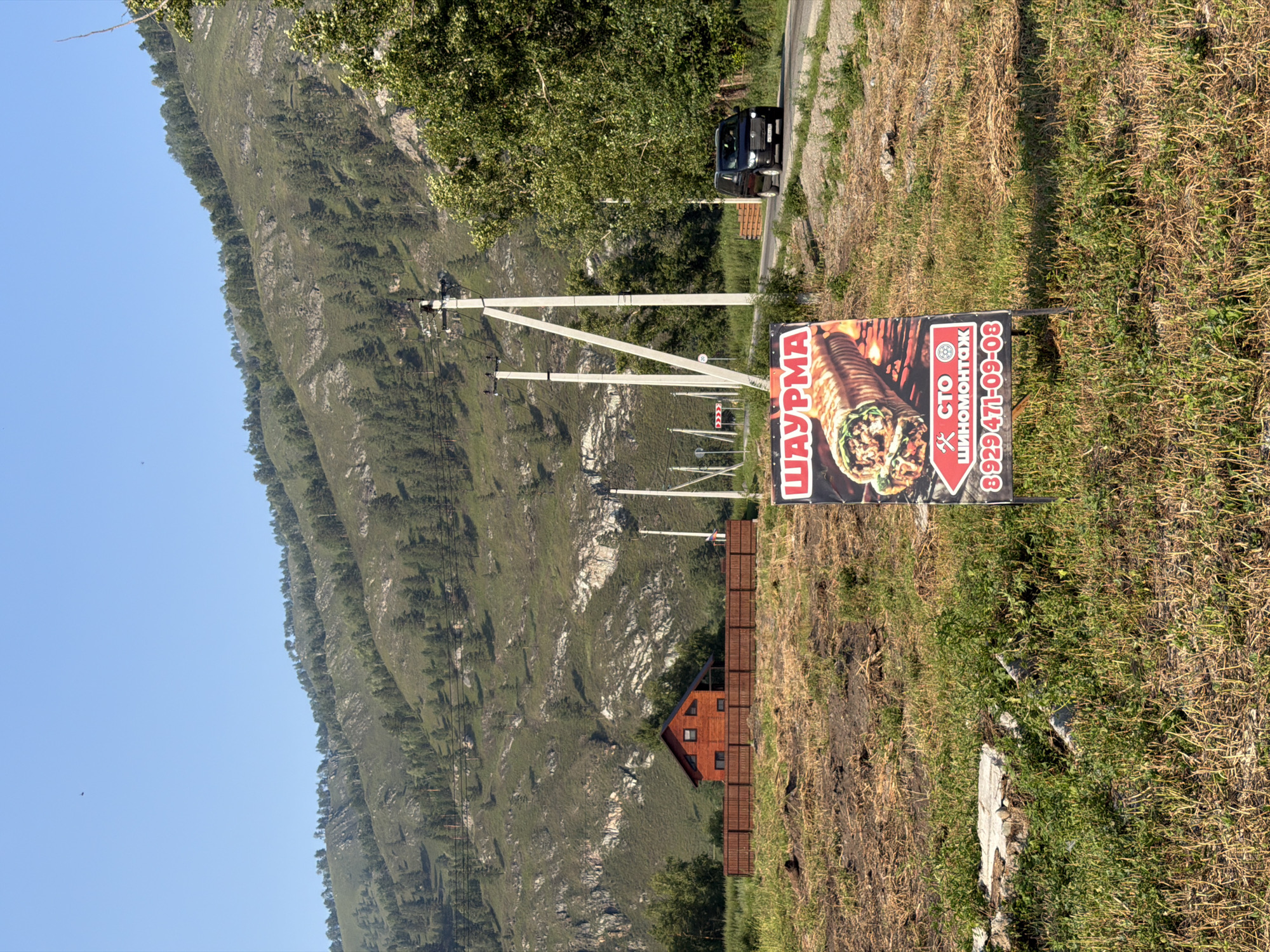 In Anos, Russia, a roadside scene unfolds with a prominent "Shawarma" sign in the foreground, featuring an image of a large shawarma wrap and a phone number. Behind the sign, a dark SUV drives along a paved road. The environment is rural, with a grassy, somewhat unkempt field leading to a wooden fence and a small, red wooden house. In the background, a large, forested mountain range dominates the landscape under a clear blue sky. Utility poles with electrical wires are visible, indicating the presence of infrastructure. The overall impression is one of a rural roadside establishment with a mountainous backdrop.