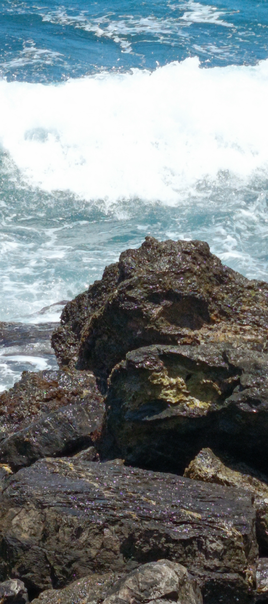 The image depicts a rocky shoreline with waves crashing against dark, wet rocks. The ocean is a vibrant blue, with white foam from the waves visible in the foreground and midground. The rocks are varied in size and texture, with some appearing smooth and rounded, while others are jagged and rough. The scene suggests a natural, untamed environment, likely along the coast of Lamkruet, Indonesia, a region known for its picturesque beaches and volcanic rock formations. No people are present in the frame, and no actions or interactions are depicted. The focus is entirely on the interplay between the water and the geological features of the coastline.