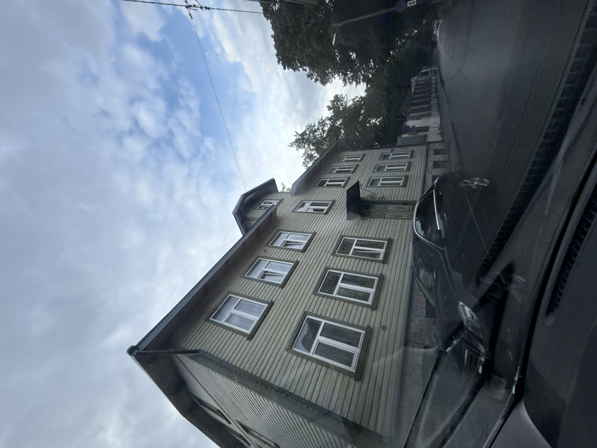 The image shows a street scene in Tallinn, Estonia, on an overcast day. A light-colored wooden building with multiple windows lines the left side of the street. A dark-colored car is parked in front of the building, and the street appears wet, suggesting recent rain. The perspective is from inside another vehicle, looking out through the windshield. Trees and power lines are visible in the background. The overall impression is a residential or commercial street in an older part of the city.