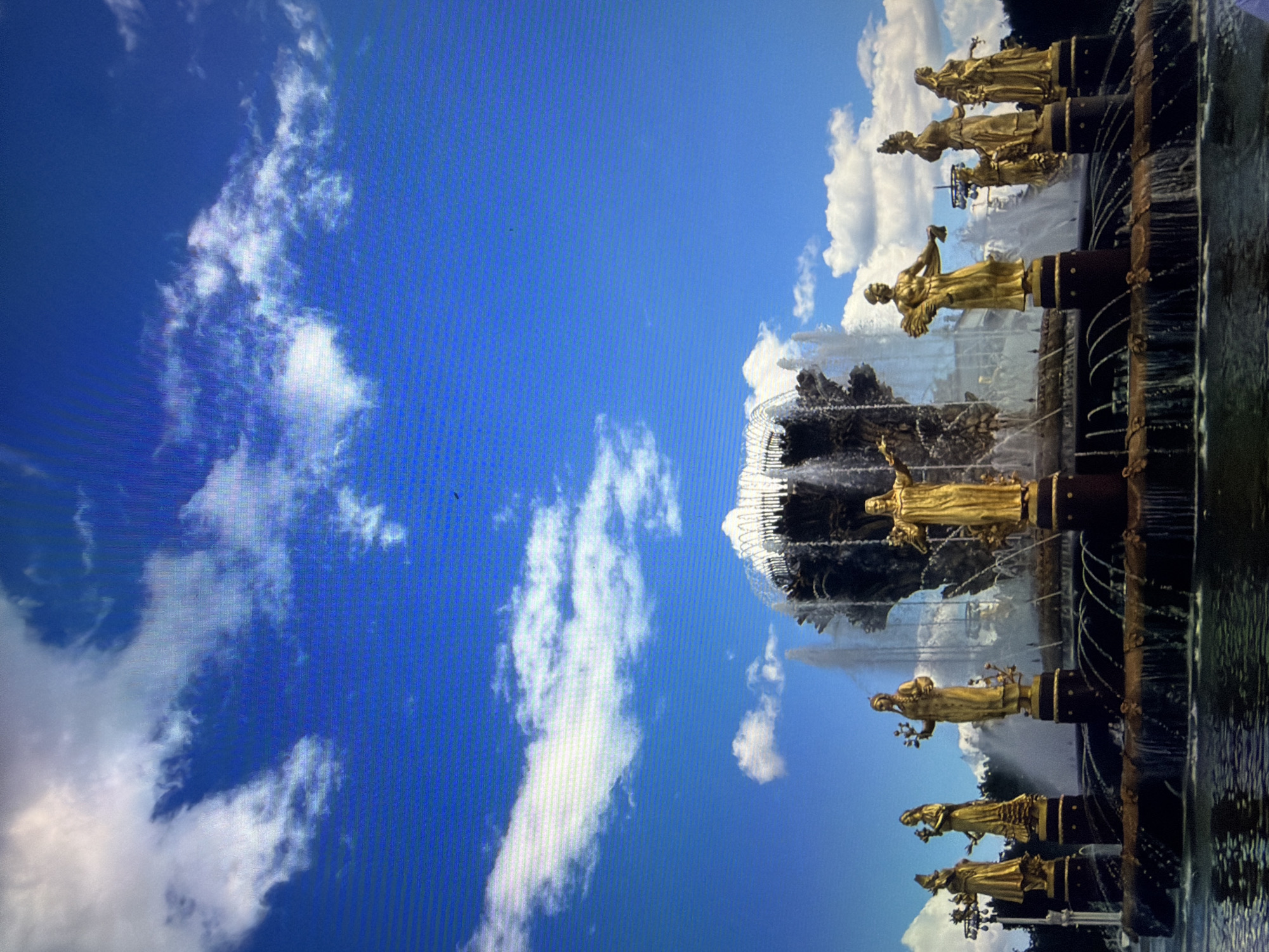 This is a full-frame, eye-level, outdoor shot of the Friendship of Nations fountain in Kotelniki, Russia. The fountain is a central focus, featuring a large tiered structure with multiple golden statues of women holding various objects, such as sheaves of wheat and flowers. Water cascades down the fountain's levels and sprays upwards, creating a dynamic display. The fountain sits in a pool of dark water, reflecting the surrounding elements. Behind the fountain, lush green trees are visible. The sky above is a vibrant blue, dotted with scattered white cumulus clouds. The overall scene conveys a sense of grandeur and artistic expression under a bright, clear sky.