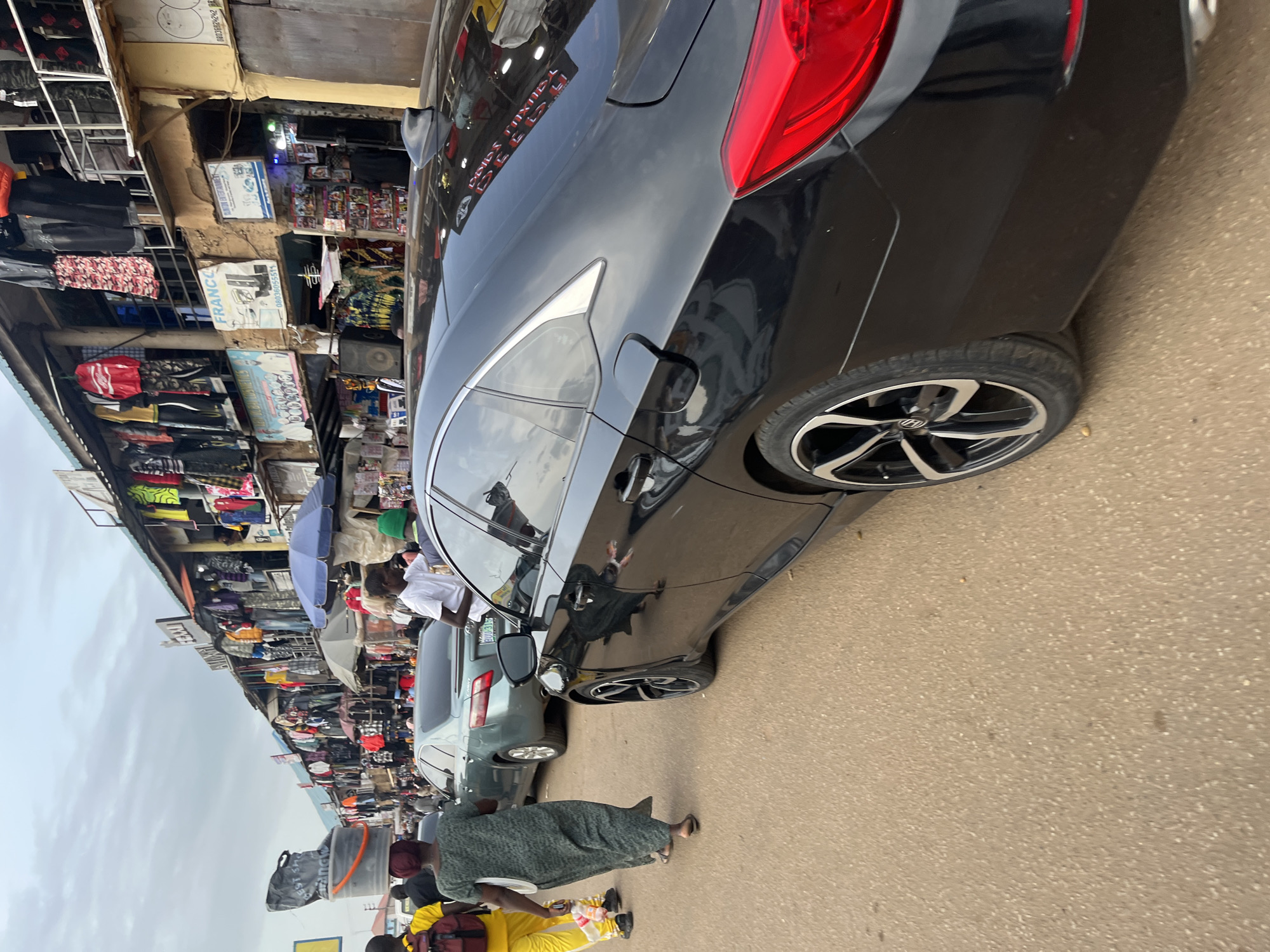 The image depicts a bustling street scene in Bukuru, Nigeria, characterized by an open-air market and heavy traffic. In the foreground, the rear section of a black sedan is prominently featured. Behind it, a silver sedan is seen, followed by a woman in a green dress carrying a large platter on her head and a man in a yellow shirt with a backpack. Further down the street, a diverse crowd of people is visible, intermingled with more vehicles. The market stalls, laden with clothing and various goods, line both sides of the street, creating a vibrant and dynamic atmosphere. The overall impression is one of a busy commercial hub.