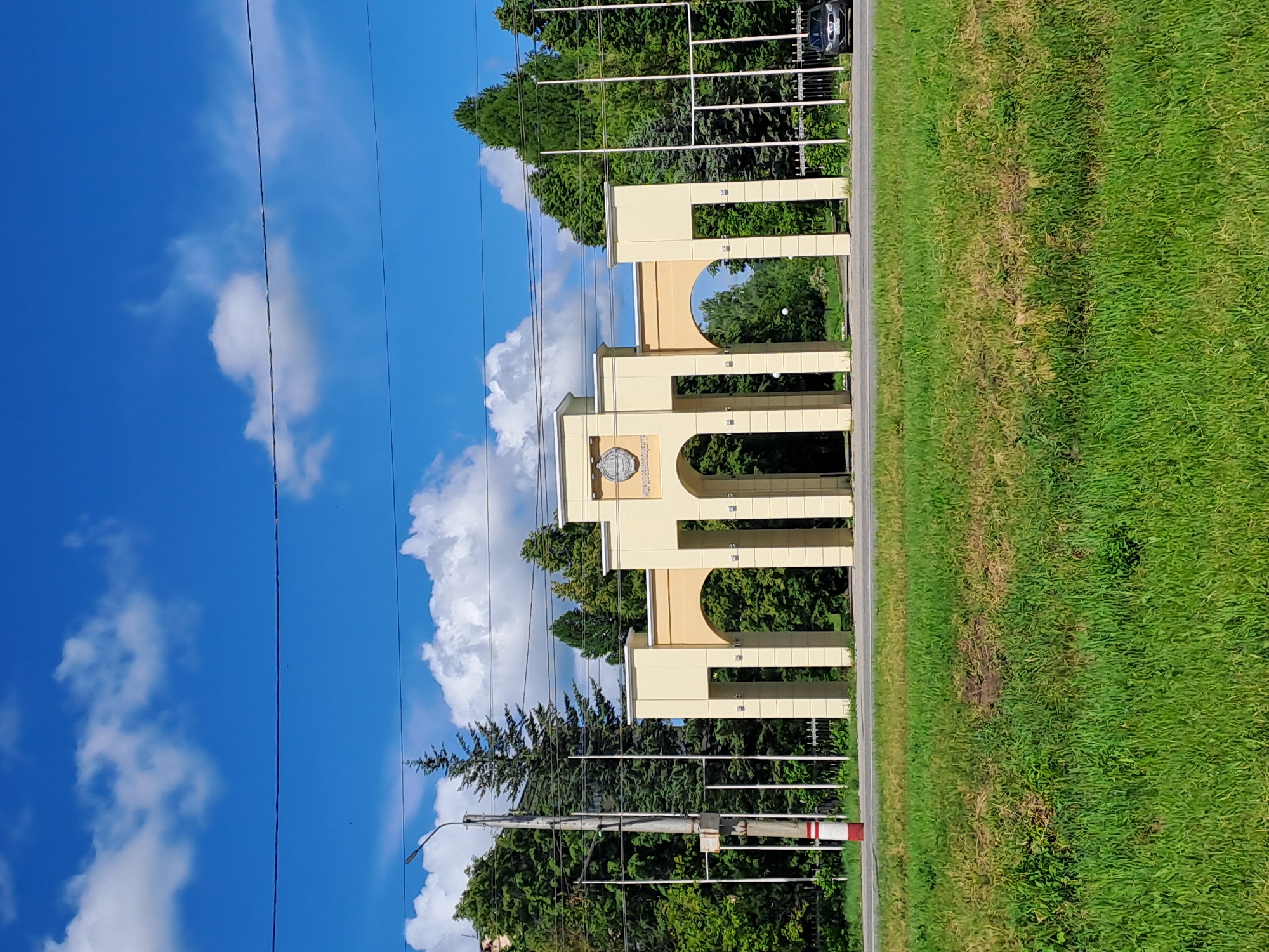 This brightly lit outdoor image, when correctly oriented, presents a scenic view of an entrance archway under a clear blue sky with scattered white clouds. The central subject is a grand, light-yellow (or beige) archway structure composed of multiple arches and pillars, likely marking an entrance to an institution. On the central part of the archway, a circular emblem or logo is visible, and beneath it, Cyrillic text reads "СЕЛЬСКОХОЗЯЙСТВЕННЫЙ ИНСТИТУТ," translating to "AGRICULTURAL INSTITUTE."
Behind the archway, a dense expanse of dark green evergreen trees forms a lush backdrop. Interspersed within the trees are several tall, thin metal structures, possibly parts of a fence or support system. To the right of the archway, a narrow paved road or path runs horizontally, separating the archway from a wide strip of vibrant green grass in the foreground, which shows some drier, lighter patches. A dark-colored car is partially visible among the trees behind the archway, suggesting a parking area or a road further back.
The time of day is clearly bright daylight, likely midday or early afternoon, with sunny weather. Several dark utility wires crisscross the blue sky, and a utility pole with a street lamp, along with a red and white striped barrier marker, is visible on the left side of the scene. The image portrays a well-maintained and institutional setting, specifically an Agricultural Institute in Yalga, Russia.