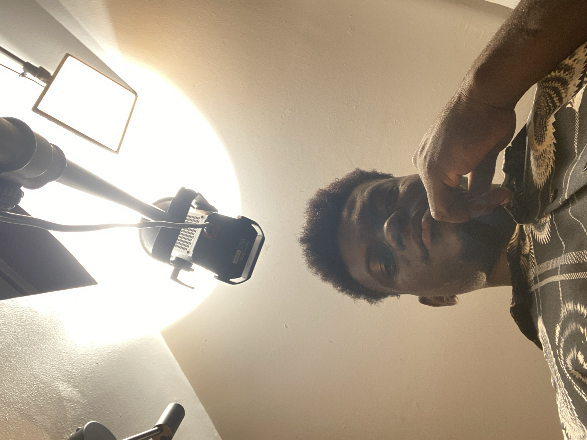 A low-angle shot looks up at a person and two bright artificial light sources against a light-colored ceiling. On the left, a rectangular LED panel or softbox emits intense white light, creating a significant lens flare. Below and to its right, another professional spotlight-style fixture with a reflector dish also glows brightly, positioned on a stand.
In the upper right portion of the frame, a person with dark skin and short, curly dark hair looks down towards the camera. Their face is partially shadowed from the upward lighting. They are wearing a dark shirt featuring a distinctive, lighter-colored patterned design, possibly geometric or abstract. One hand is visible near their neck or chest area, appearing to be adjusting something or part of a pose. The strong, controlled lighting setup suggests an indoor professional setting, such as a studio, likely for a photoshoot or video recording. No text is visible in the image.