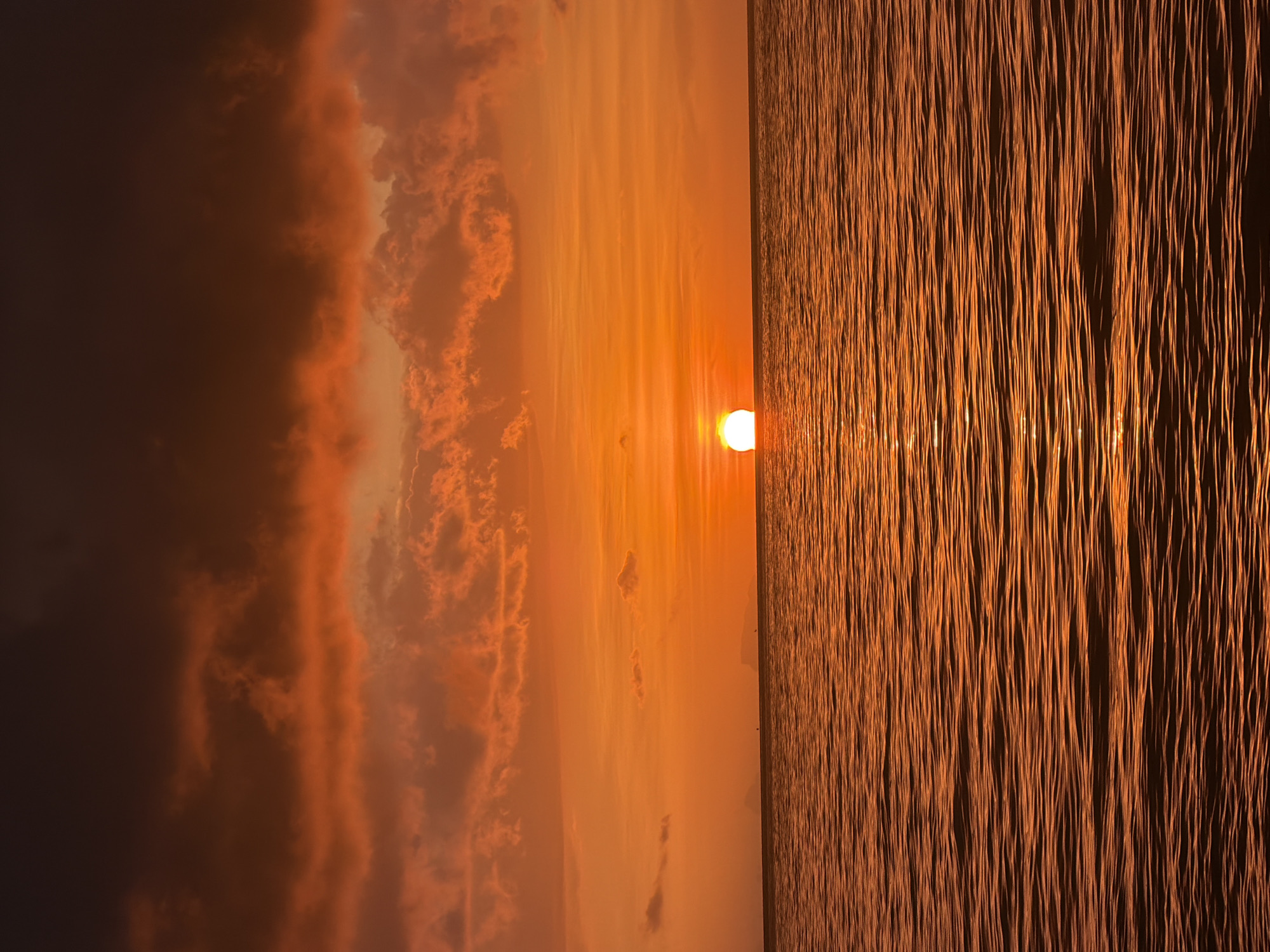 A sunset over the ocean at Baan Tai, Thailand, features a vibrant orange sky filled with dramatic clouds. The sun, a bright orange orb, is setting on the horizon, casting a shimmering reflection on the water's surface. The ocean is calm, with gentle ripples creating a textured pattern. In the distance, faint silhouettes of landmasses can be seen. The overall scene is one of serene natural beauty.