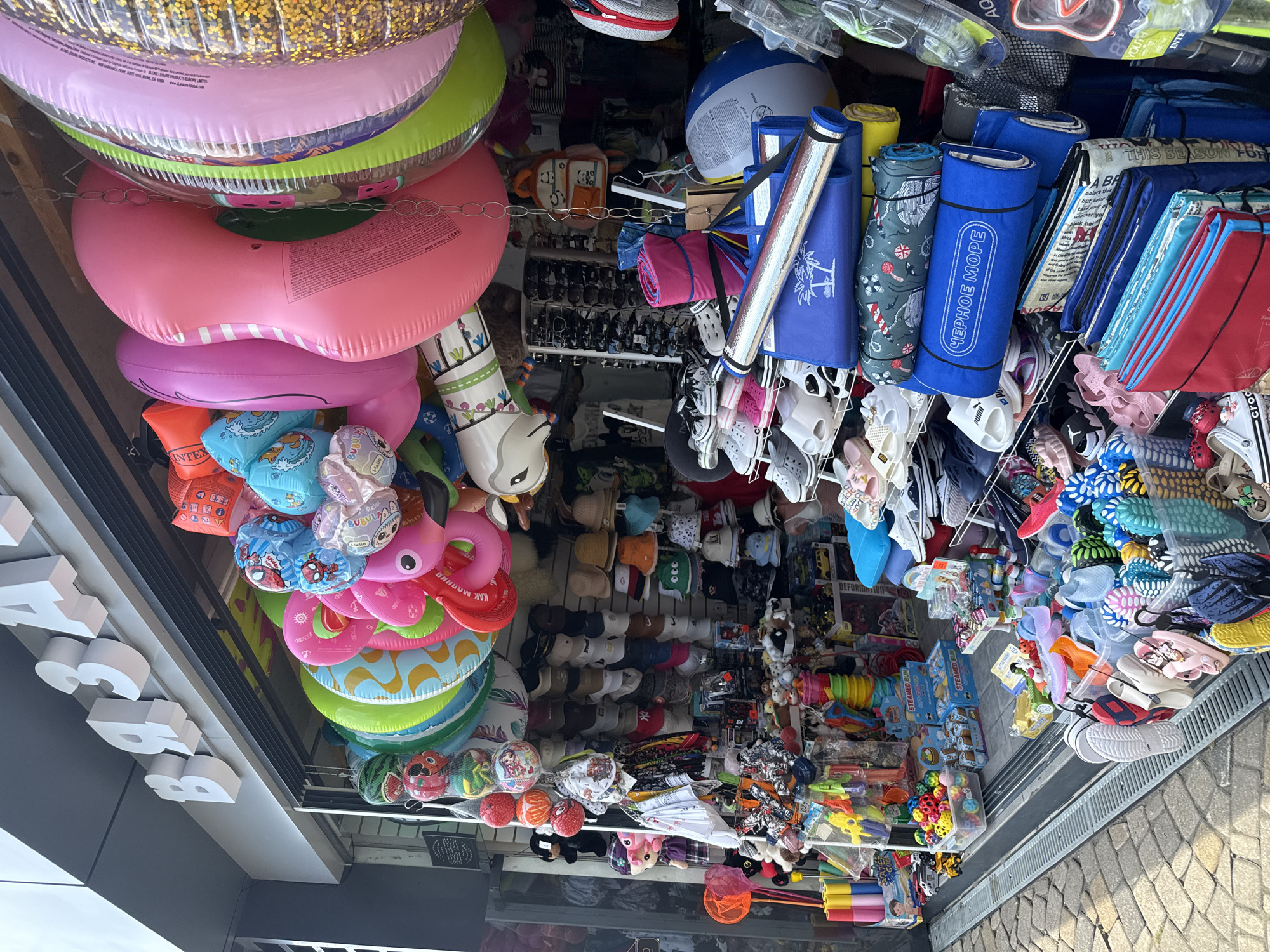 The image depicts a vibrant street-level shop stall in Sochi, Russia, laden with a diverse array of summer merchandise. Prominently displayed at the top are inflatable rings and pool floats, including a large pink flamingo, a watermelon-themed ring, and a ring with a glittery gold interior. Below these, a tiered display showcases an assortment of hats, sunglasses, and bags. The lower sections are filled with footwear, such as flip-flops and sandals in various colors and designs, as well as children's toys like squirt guns and balls. Some of the merchandise includes branded items and children's character themes like Spider-Man and Winnie the Pooh. The stall is situated on a paved walkway, suggesting a bustling tourist area or promenade. The overall impression is one of a commercial establishment catering to beach and recreational needs.