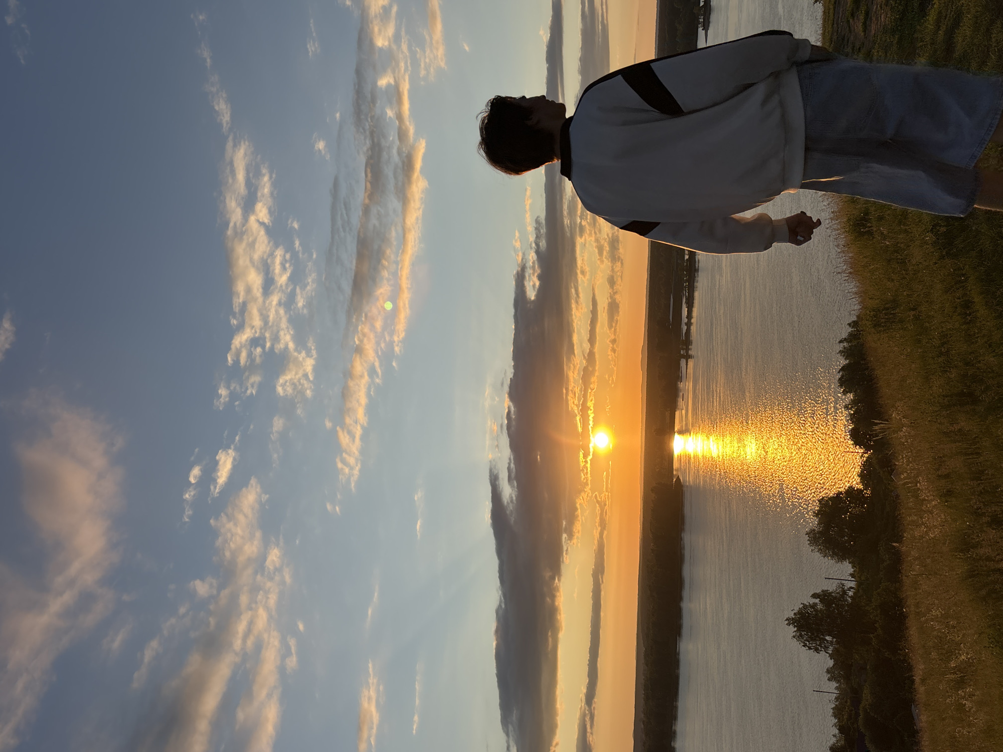 A person stands on a grassy hill overlooking a wide river at sunset. The sky is a blend of blue, orange, and pink, with scattered clouds. The sun, a bright orange orb, is low on the horizon, casting a golden reflection on the water. The person, seen from behind, is wearing a white sweatshirt with dark stripes on the shoulders and light-colored shorts. The hill is covered in dry grass and scattered trees. The river is calm, with a tree-lined shore visible in the distance. This scene likely takes place in the vicinity of Olkhovka, Russia, given the geographical context often associated with such riverine landscapes.