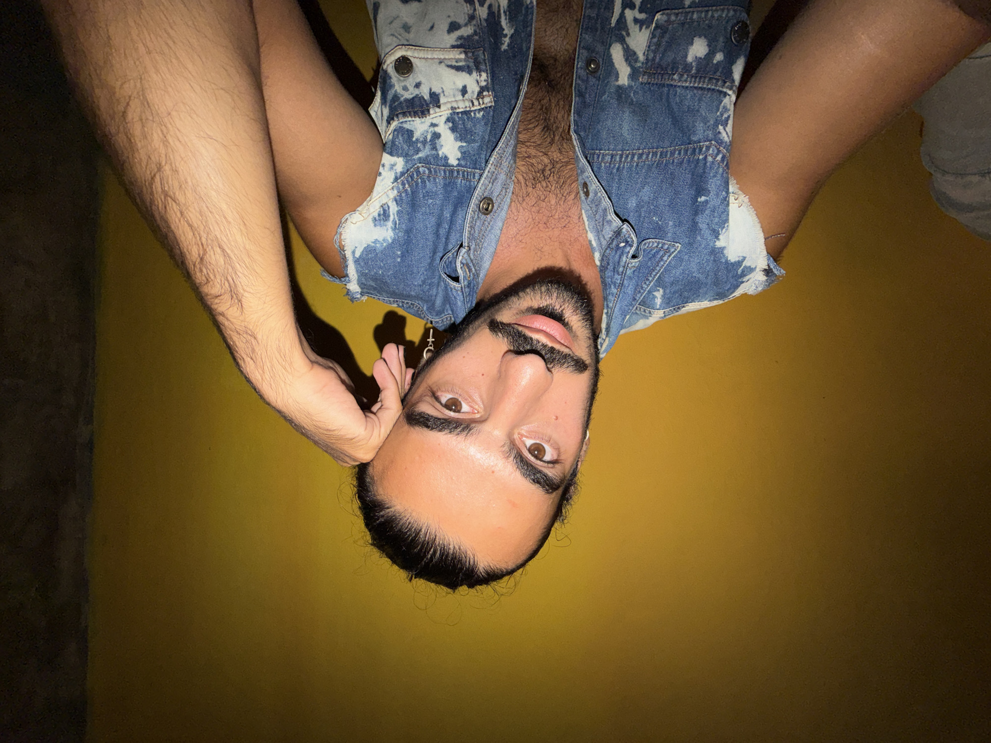A man with dark hair pulled back into a bun, a mustache, and a beard is leaning his head on his hand, looking at the camera. He is wearing a sleeveless denim vest with white bleach spots over a dark shirt that is unbuttoned, revealing dark chest hair. He has a cross earring in his left ear. The background is a bright yellow wall. The image appears to be taken in North Kuta, Indonesia, likely indoors due to the lighting and the solid yellow wall.