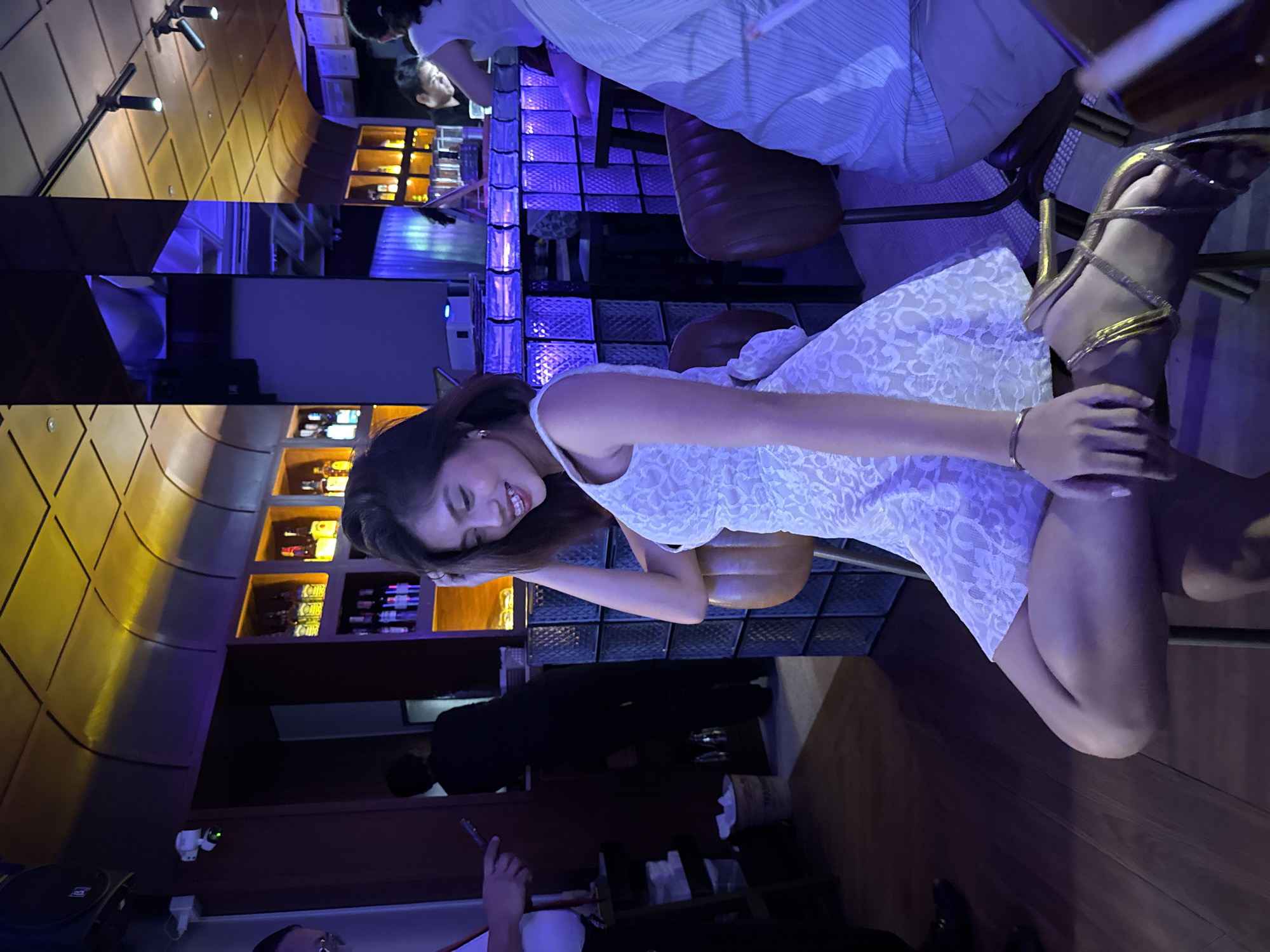 In a dimly lit establishment with ambient lighting, a woman in a white lace dress sits on a brown chair. She has her hand on her hair and is smiling. She is wearing gold strappy high heels. The background shows a bar area with bottles on shelves, a counter with blue lighting, and other patrons. The scene appears to be a bar or lounge in Davao City, Philippines, suggested by the presence of decorative elements that are common in such establishments in the Philippines.