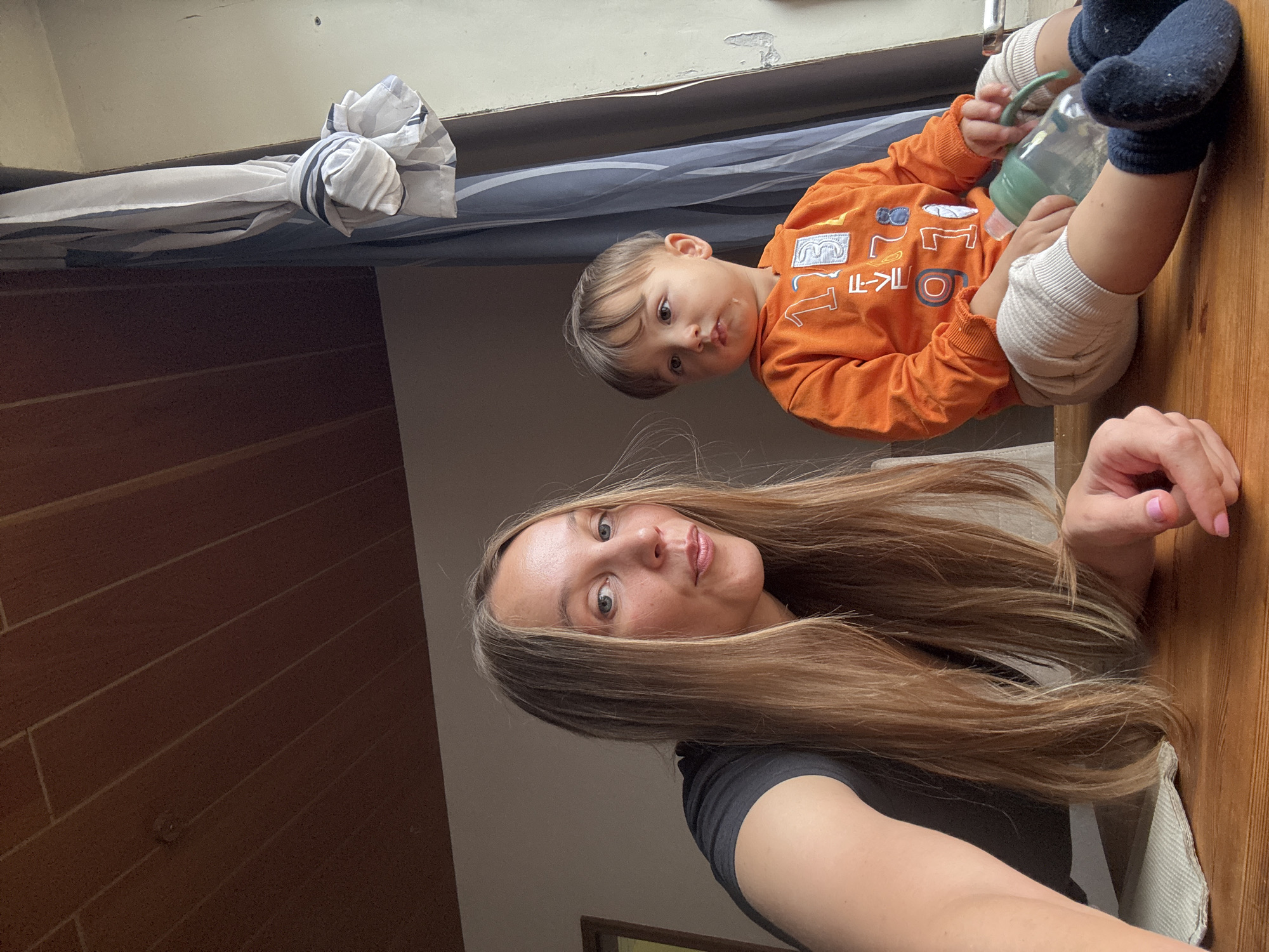 The image depicts a woman and a toddler sitting at a wooden table. The woman, with long blonde hair and wearing a black t-shirt, is on the left side of the frame, looking towards the camera. The toddler, on the right, is wearing an orange long-sleeved shirt with colorful numbers and white ribbed leggings. The toddler is holding a green sippy cup. In the background, a window with a draped curtain is visible. The environment appears to be an indoor setting, possibly a home. There is no discernible indication of the specific location within Überherrn, Germany.