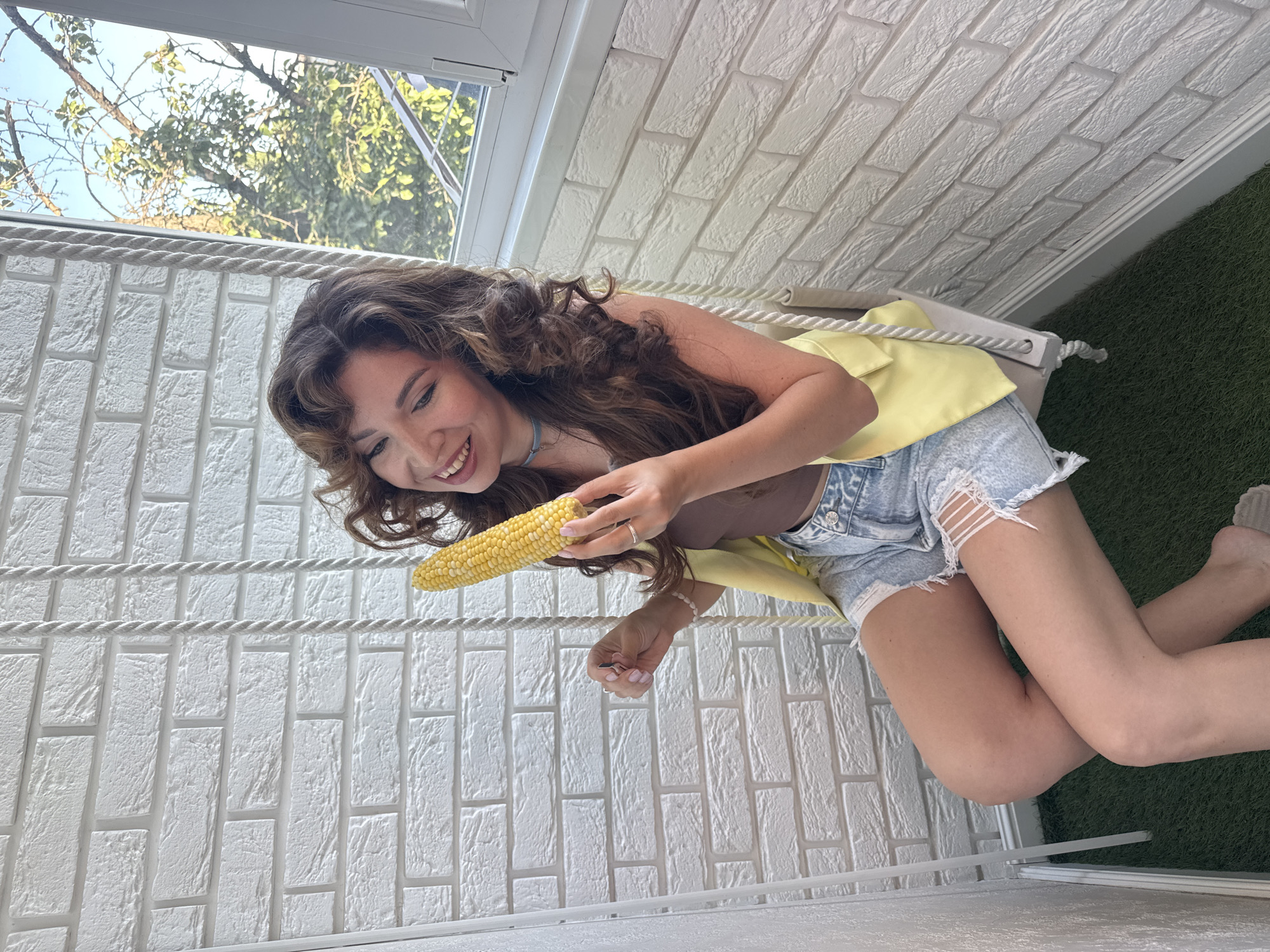 A young woman with curly brown hair is seated on a white swing, holding a cob of corn. She is wearing a brown top, a yellow vest, and distressed denim shorts. The background features a white brick wall and a window with green foliage visible outside. The floor is covered in green artificial turf. The scene is indoors, likely on a balcony or enclosed porch, and appears to be in Kryvyi Rih, Ukraine.