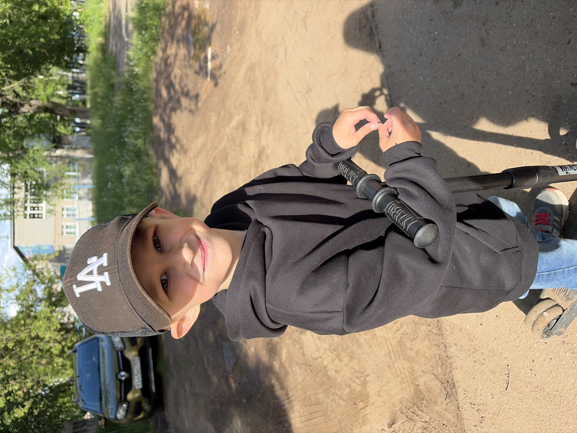 A young boy wearing a black hoodie and a black baseball cap with the "LA" logo is standing on a dirt path in the city of Chaikovsky, Russia. He is holding the handlebars of a scooter. He is smiling at the camera. A yellow car is visible in the background, parked near trees and a building.