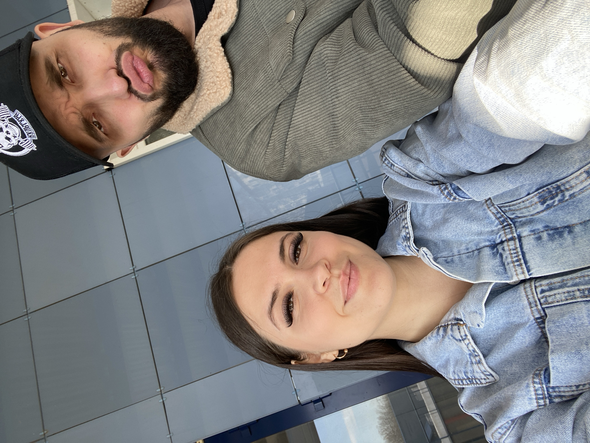 This is a portrait-style photograph featuring a man and a woman, taken in Chaikovsky, Russia. The image is oriented sideways, with subjects looking towards the camera.
The man, positioned in the upper left of the frame (when viewed upright), has a dark beard and is wearing a black baseball cap with a white circular logo featuring text, which appears to include "BARTYHA" or similar stylized letters. He is also wearing a grey corduroy jacket with a cream-colored sherpa lining. He is making a playful expression with pursed lips and slightly squinted eyes.
The woman, positioned in the lower right, has long dark hair and is wearing a light blue denim jacket over a lighter-colored top. She has noticeable long eyelashes and small gold hoop earrings. She is smiling softly at the camera with a calm expression.
The background consists of a blue tiled or paneled wall, suggesting an outdoor or brightly lit indoor setting. The lighting is bright and even, indicating daytime, possibly sunny weather. The overall mood is casual and pleasant.