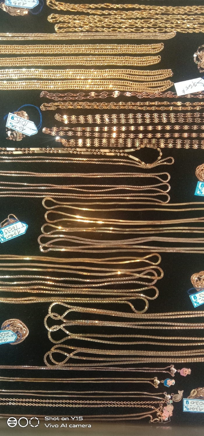 This image depicts an overhead, close-up view of a retail jewelry display in Maron, Indonesia. The scene features a collection of approximately 40 distinct gold-colored chains, presented on a dark, non-reflective surface, likely a velvet or similar material display mat. The chains vary in link style, including curb, Figaro, snake, box, and flat-disc designs. They are primarily arranged in parallel rows, with some chains coiled or looped. Several small, light-colored rectangular price tags, some attached with blue string, are visible among the jewelry, displaying handwritten or printed numerical values and percentages such as "42.5%", "695.K", and "1.070". Some chains are adorned with small, colored charms resembling cartoon figures, animals, or anchors. A few coiled rings, matching the gold color of the chains, are also present. The metallic surfaces of the jewelry exhibit multiple reflections from an overhead light source. No individuals or active interactions are depicted.