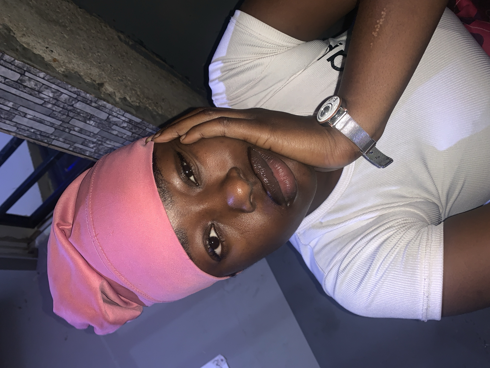 A dark-skinned individual is featured in a close-up shot. They are wearing a pink head covering and a white ribbed top. A silver watch is on their left wrist. The person's left hand is positioned on their forehead. The background includes what appears to be a wall with decorative tiles. The location is New Stadium, Nigeria.