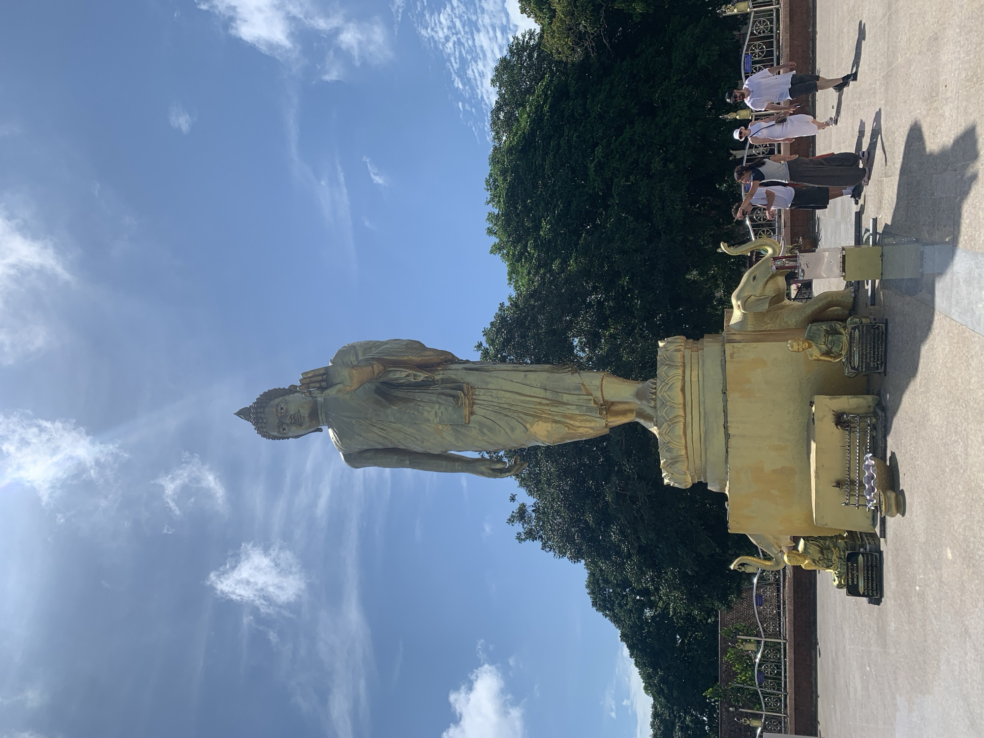 A golden statue of Buddha stands tall against a bright blue sky dotted with white clouds. The Buddha is in a standing pose, with its right hand raised in a gesture of blessing or protection. The statue is placed on a large golden pedestal. In front of the pedestal, several smaller golden statues of Buddha are visible, along with decorative golden elephants. Some people are walking in the foreground, observing the statues. The background features lush green trees and a fence. This scene appears to be at a temple or religious site on the island of Ko Samui, Thailand.