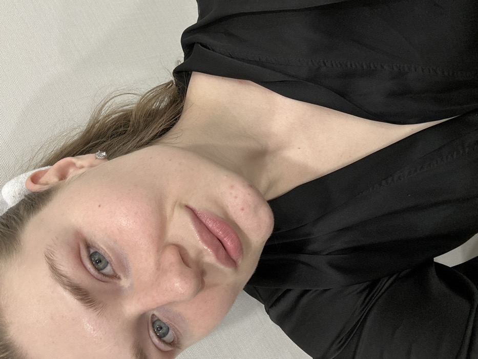 A fair-skinned woman with light blue eyes looks at the camera. She has visible pores, pink lips, and a small mole on her left cheek. She wears a black top and a white headband. An earring sparkles in her left ear. The background appears to be a light gray or white textured surface. The image is rotated slightly counter-clockwise.
